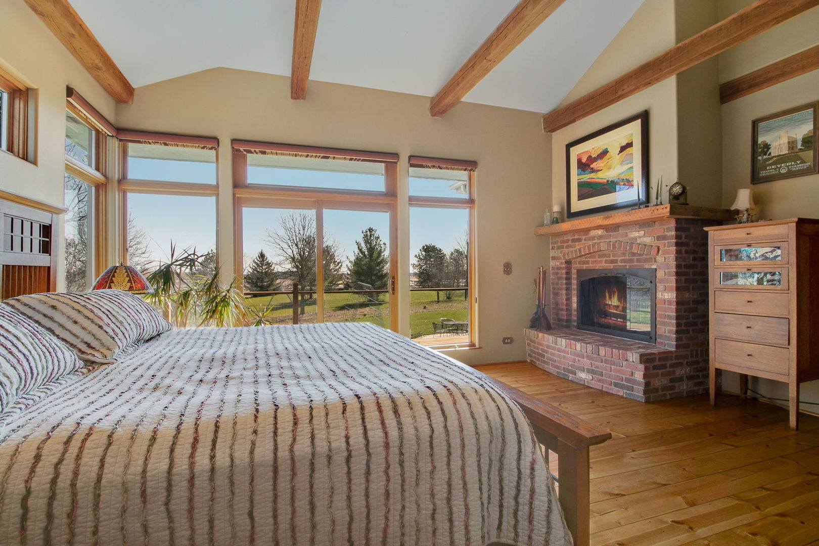 Bedroom with exposed wood ceiling beams, a brick fireplace, and tall windows overlooking a lawn.
