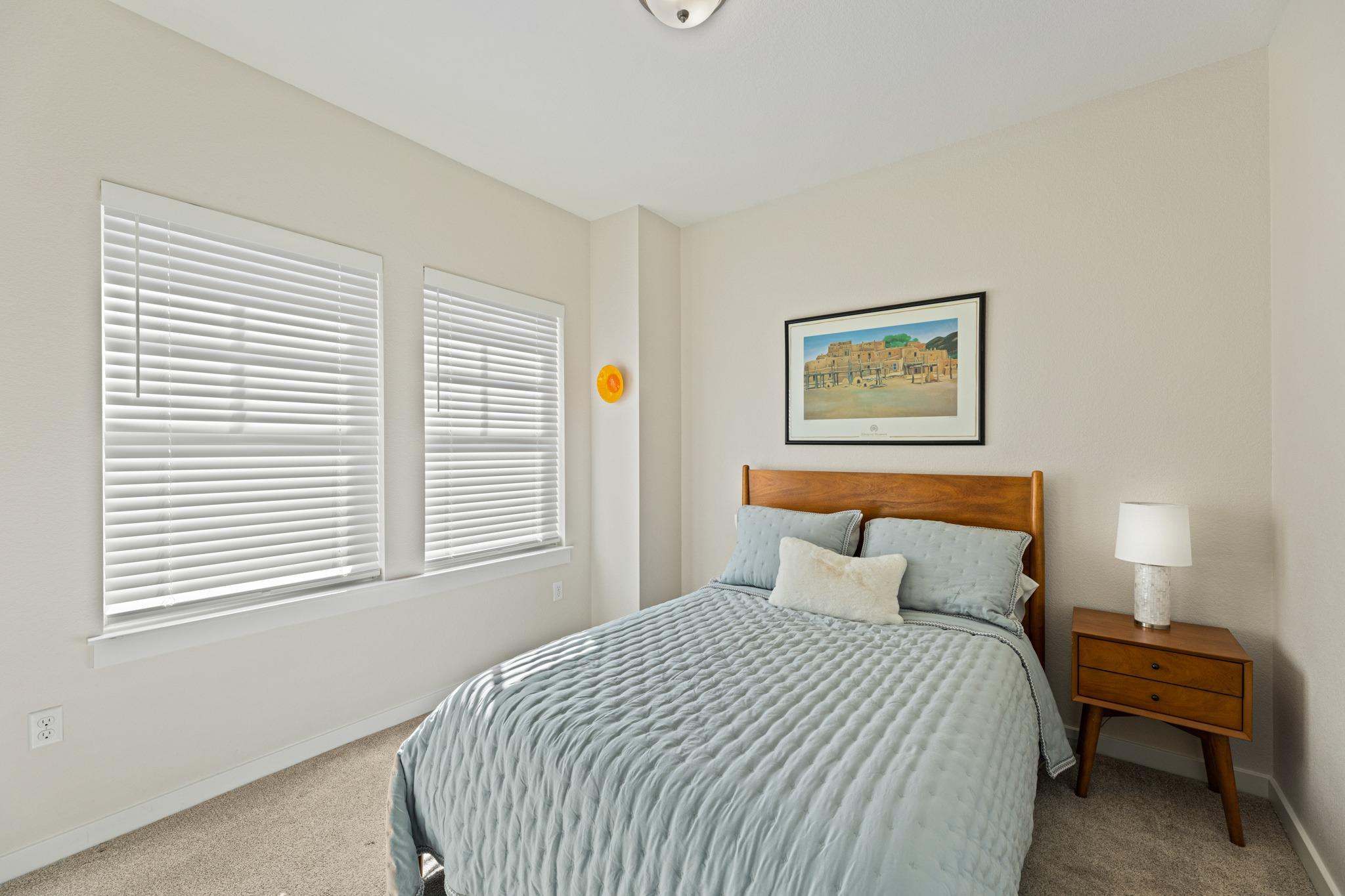 Bedroom with pale blue bedding, a wooden bed, and two windows with white blinds.