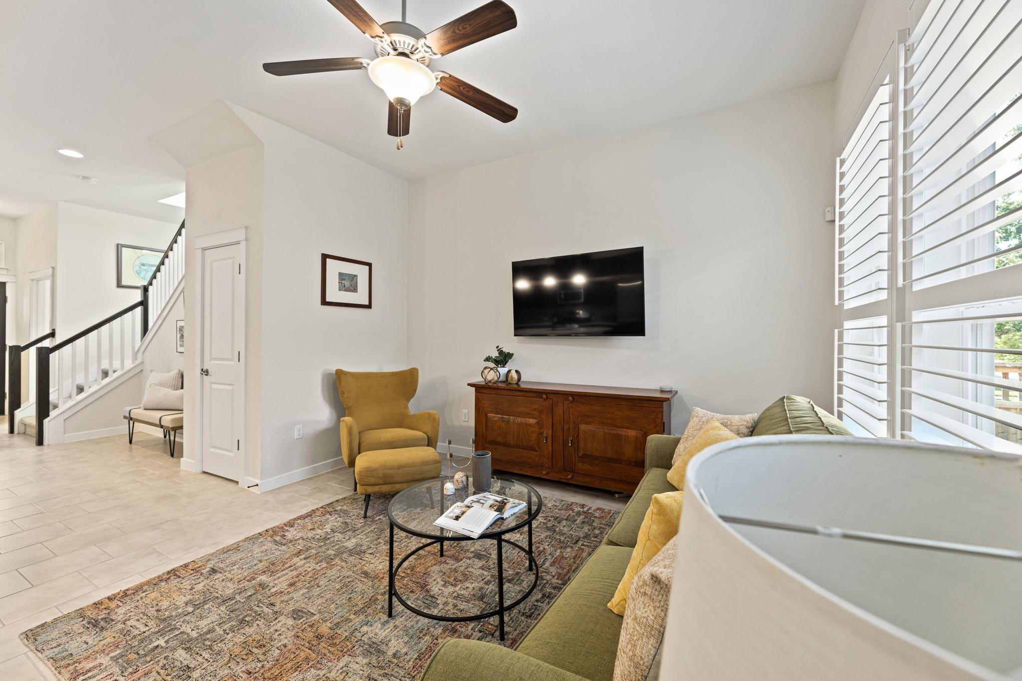 Living room with a green sofa, mustard chair, wall-mounted TV, and large plantation shutters.