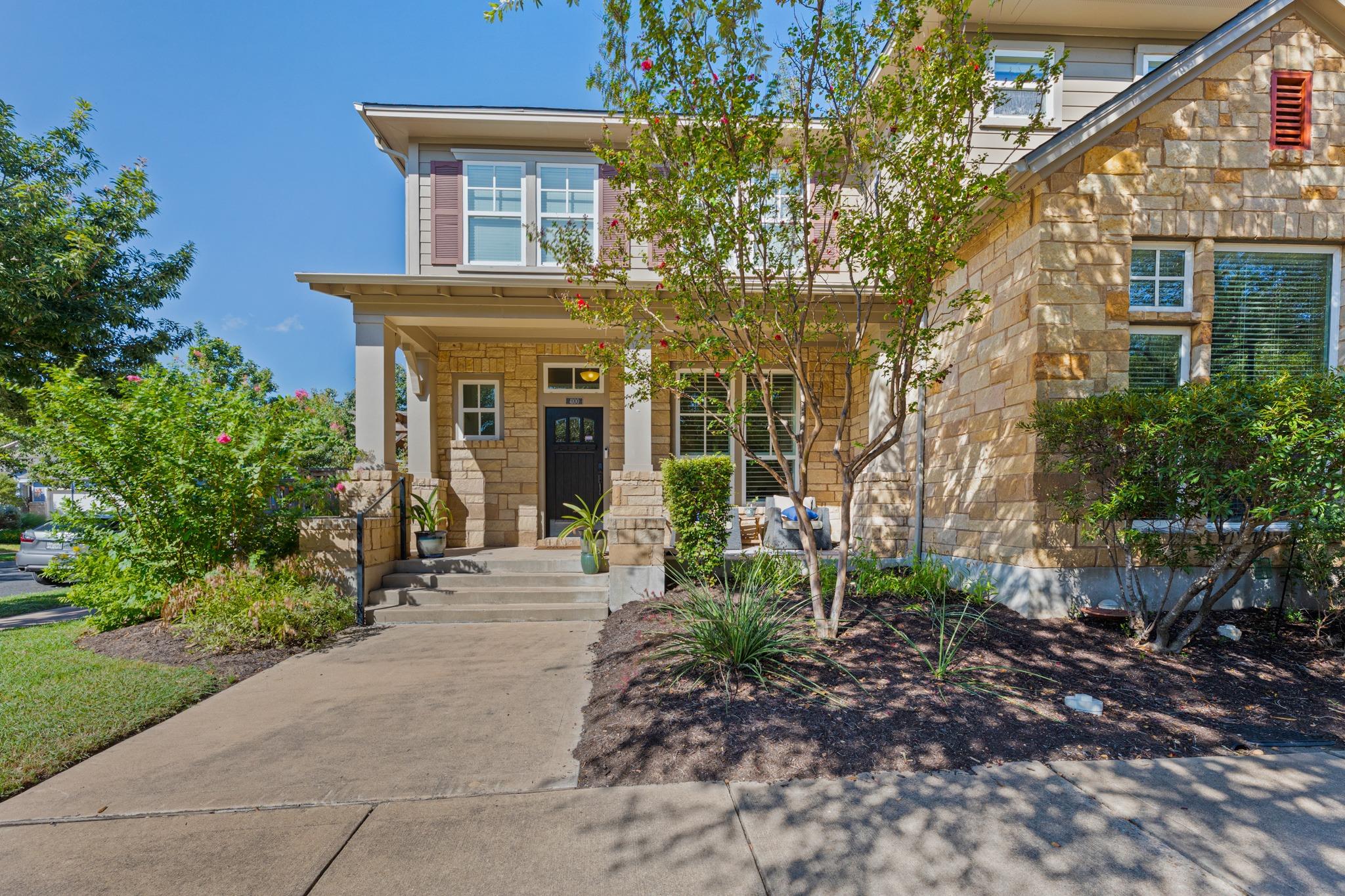 Two-story stone house with a covered entry, concrete walkway, and flowering trees.