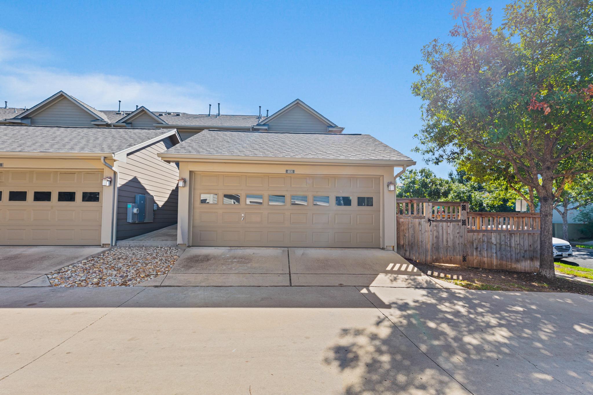 Attached home with a tan double garage, concrete driveway, wooden fence, and large shade tree.