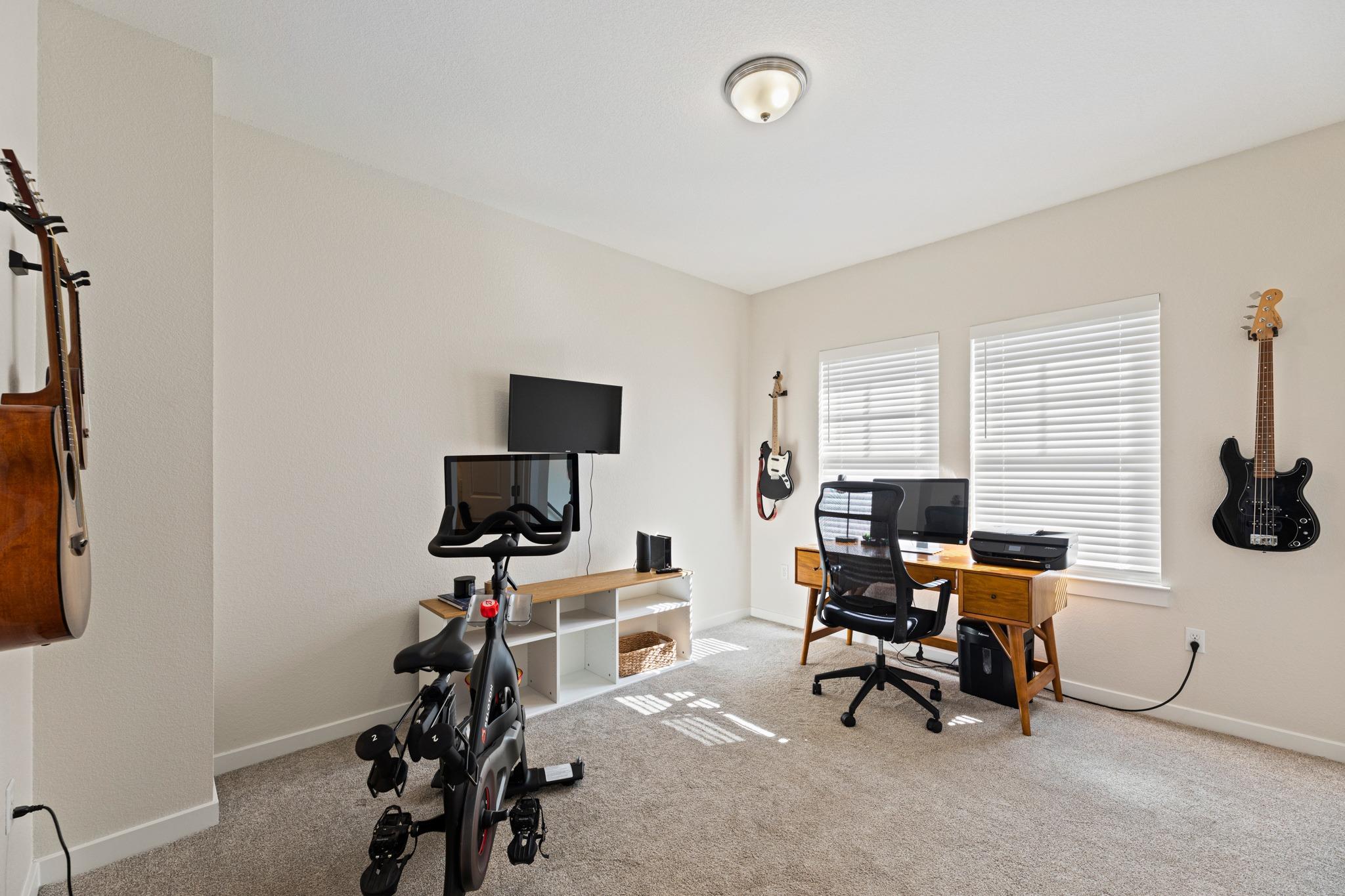Home office and workout room with a desk, exercise bike, dumbbells, and guitars on the walls.
