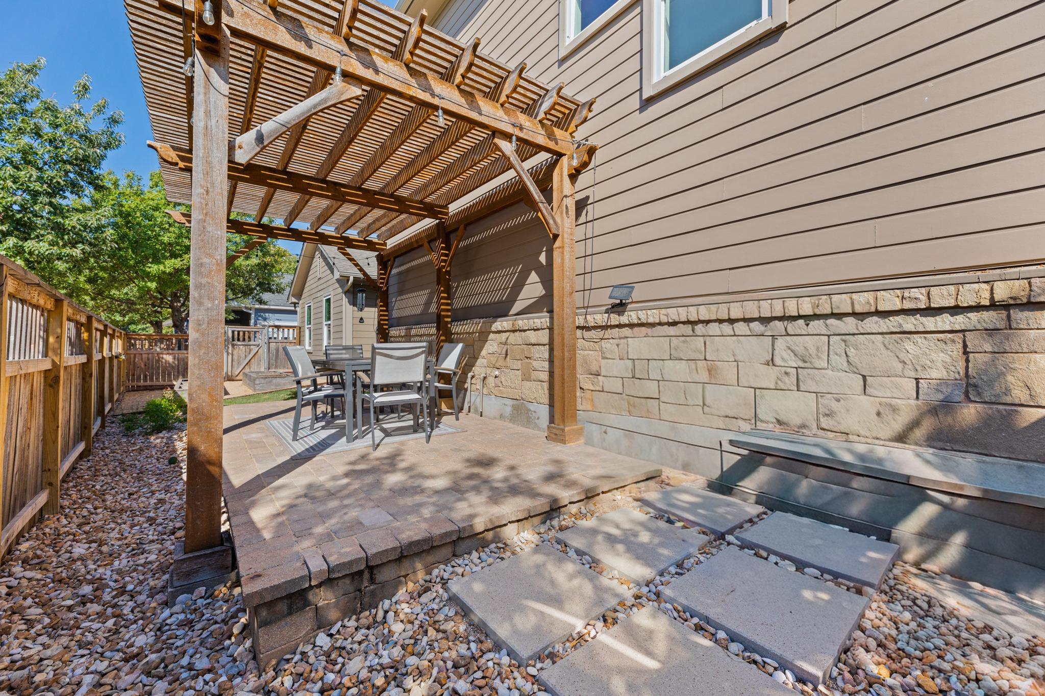 Backyard patio with a dining set beneath a wooden pergola and stepping stones across gravel.