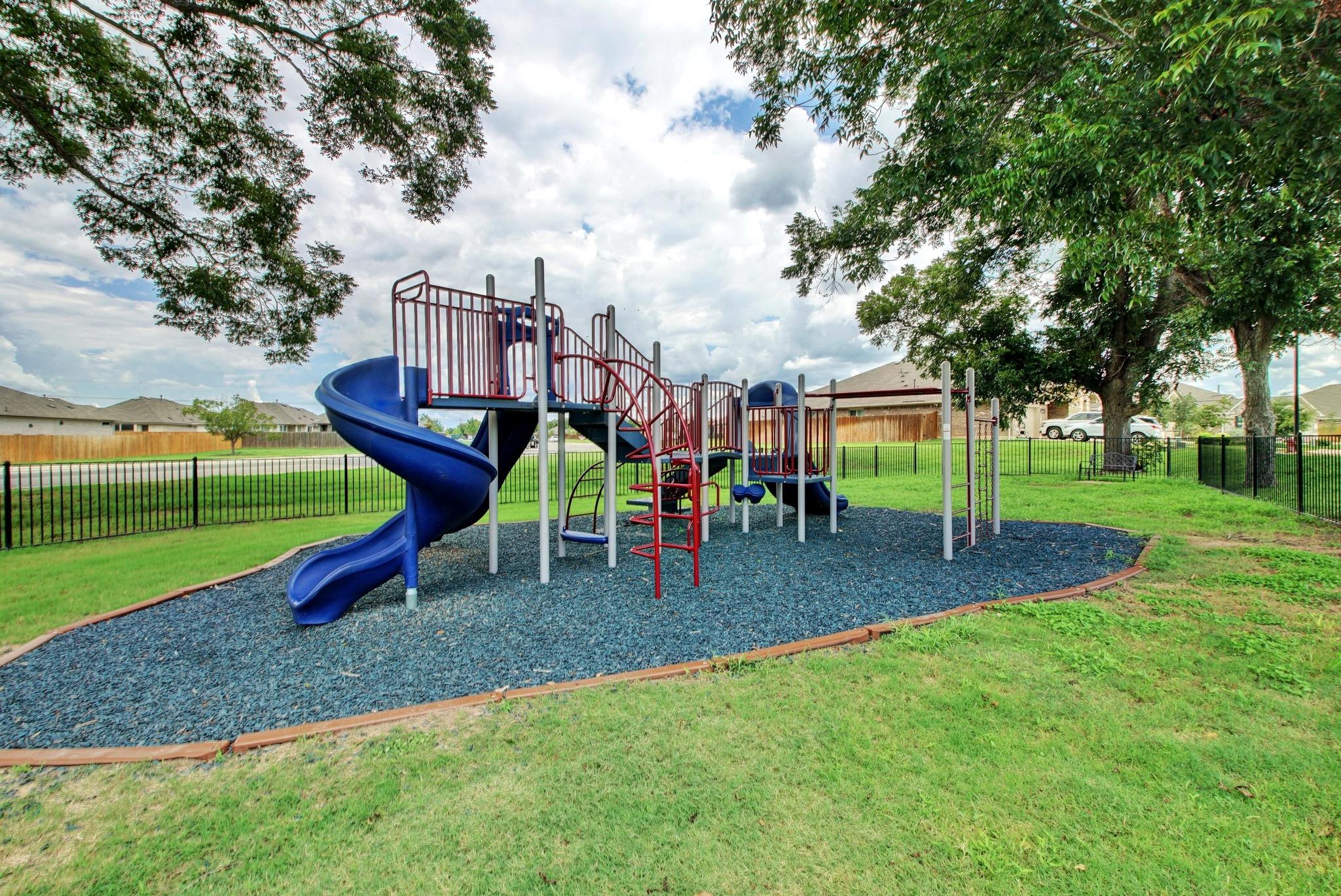 Neighborhood playground with a blue spiral slide, climbing structures, mature trees, and fenced grassy grounds.