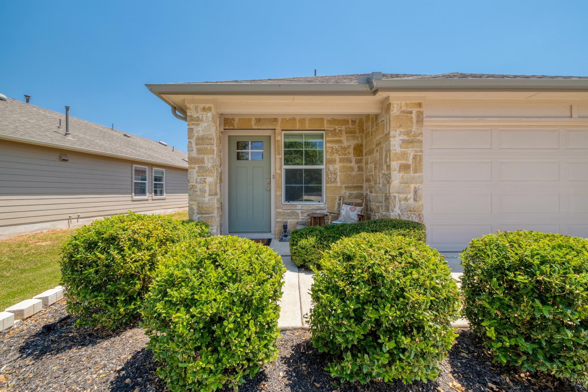 Stone-front home with a green entry door, attached garage, and trimmed shrubs lining the walkway.