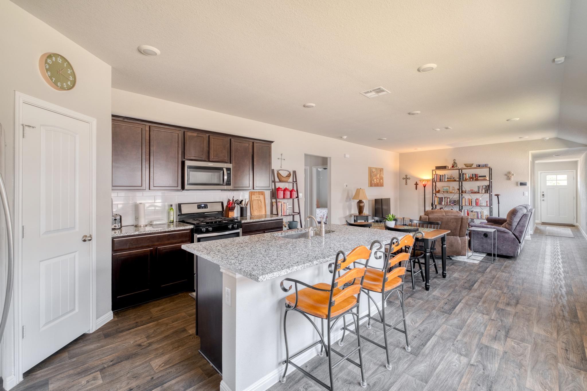 Open-plan kitchen and living room with a granite island, bar stools, dark cabinets, and a furnished seating area.