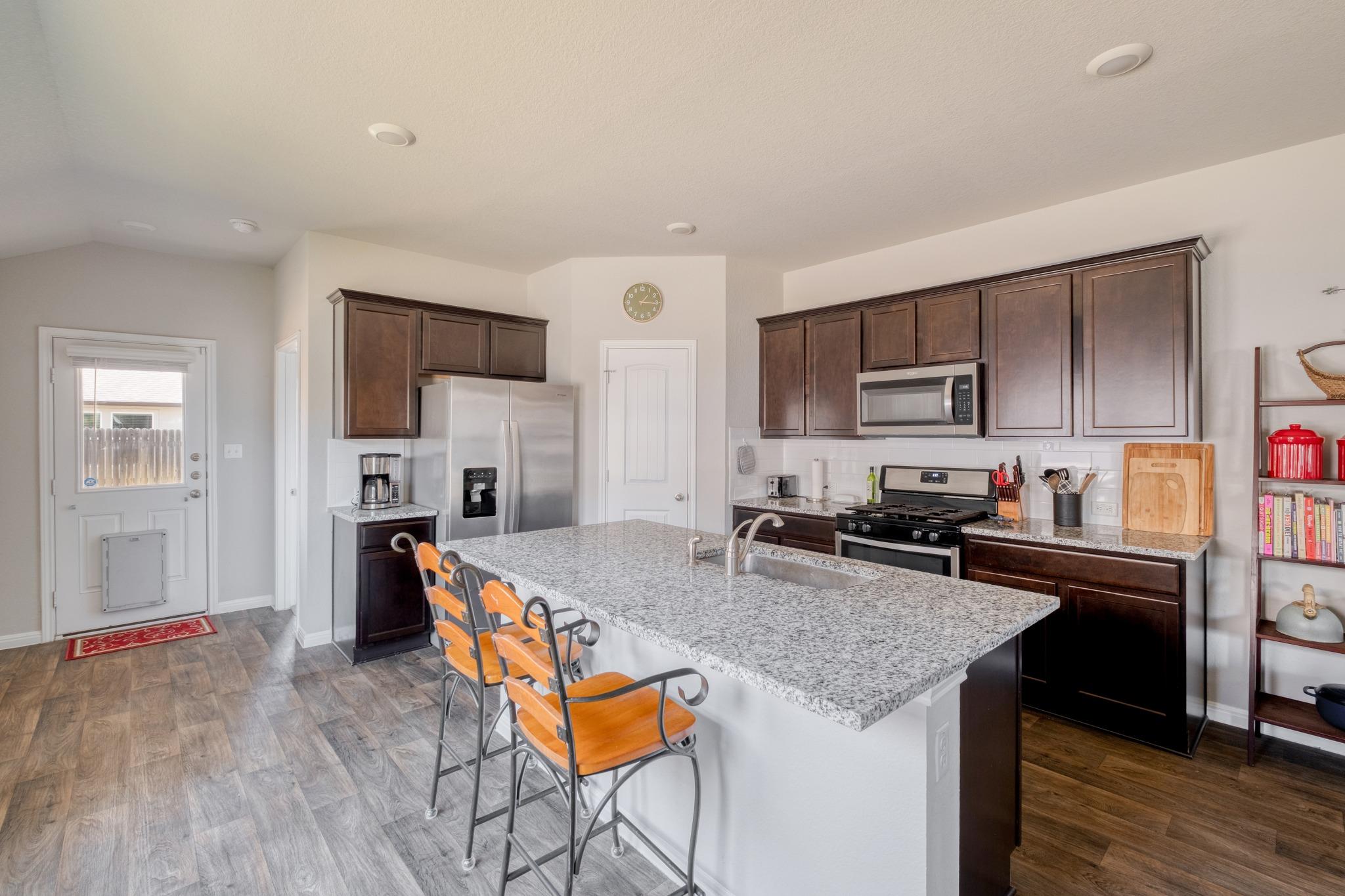 Kitchen with a large granite island, dark wood cabinets, stainless appliances, and orange bar stools.