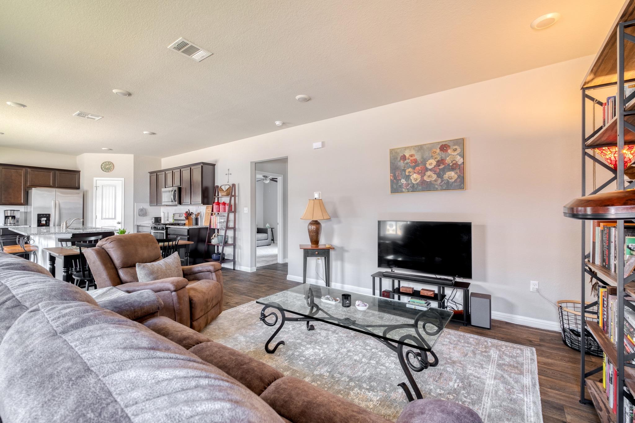 Open-plan living room with brown reclining sofas, glass coffee table, and kitchen with dark cabinets.