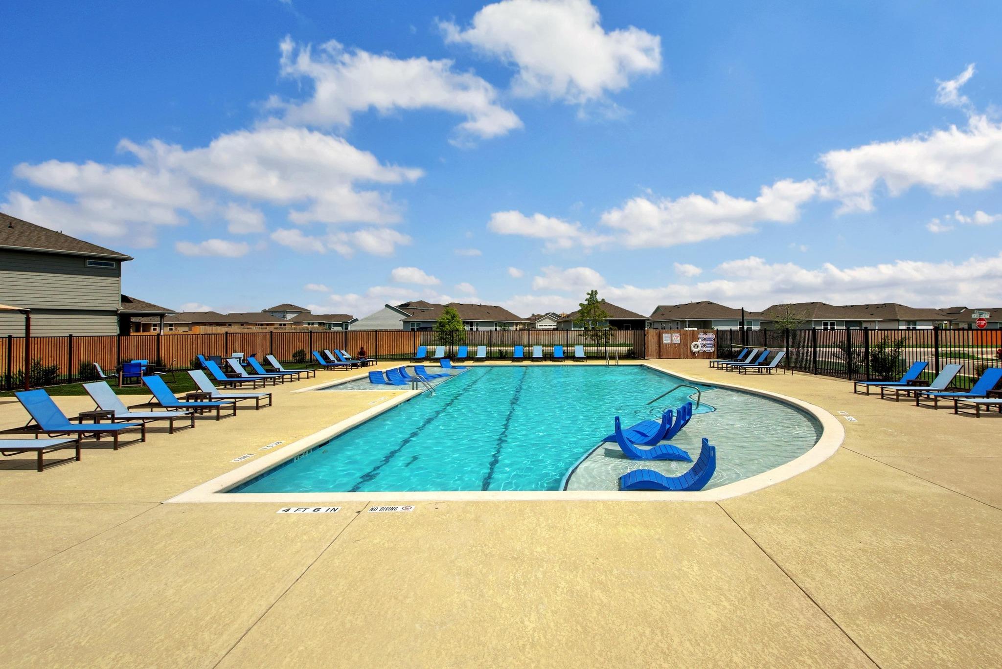 Community pool with blue in-water loungers and poolside chairs, enclosed by fencing.