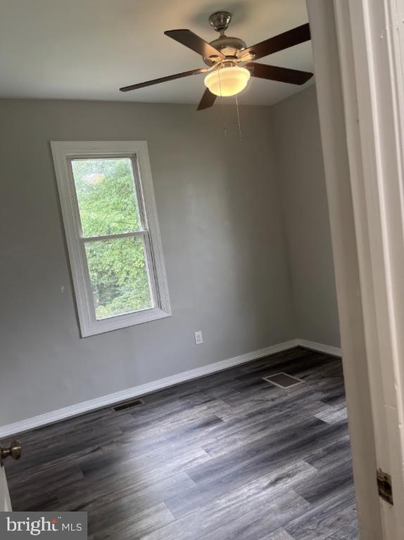 Empty room with gray walls, dark flooring, a window, and a ceiling fan with light.