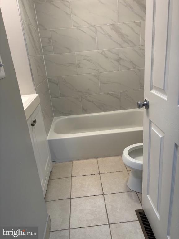 Bathroom with a white tub, gray tiled surround, beige tile floor, and toilet.