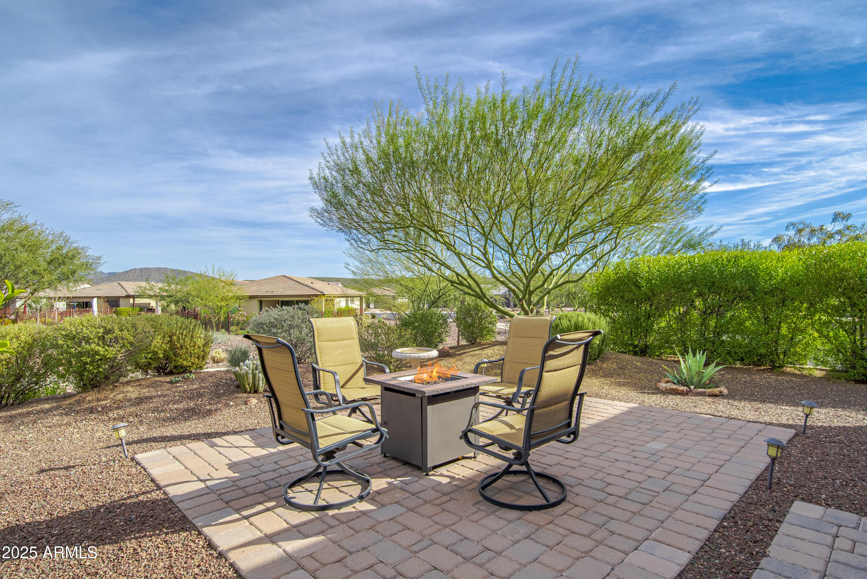 Backyard paver patio with four chairs around a lit fire pit beneath a large leafy tree.