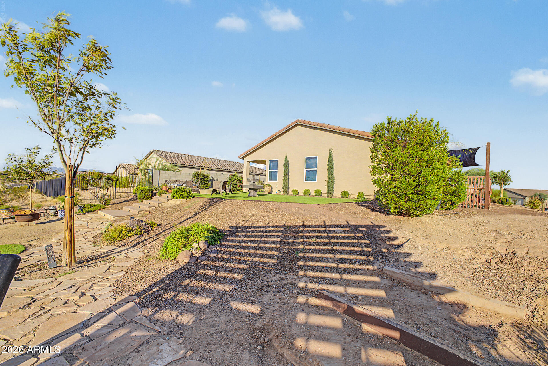 Backyard with a stone patio, gravel landscaping, outdoor seating, and a shade sail beside a tan stucco home.