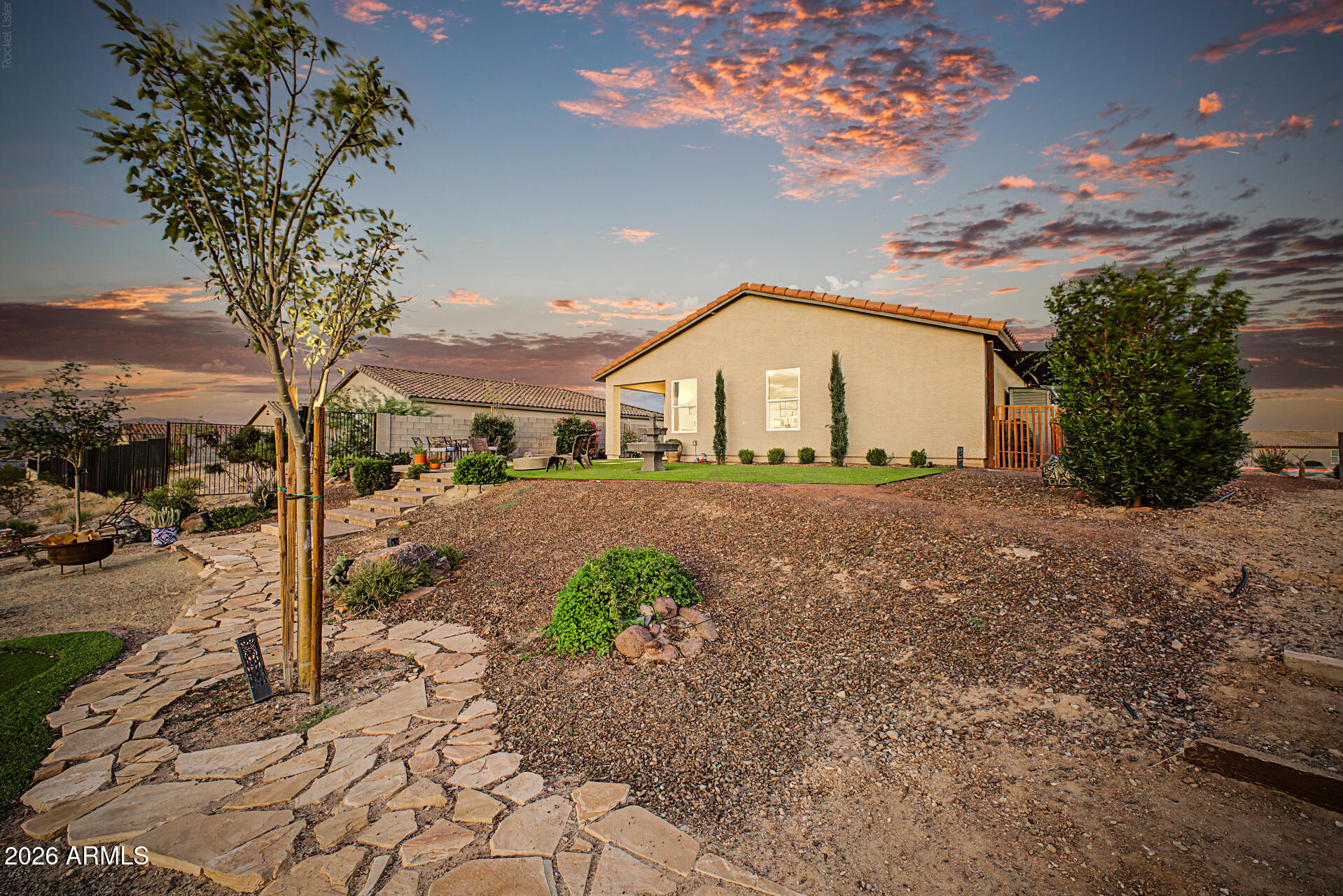 Home's rear exterior at dusk, with a gravel yard, stone path, and patio seating.