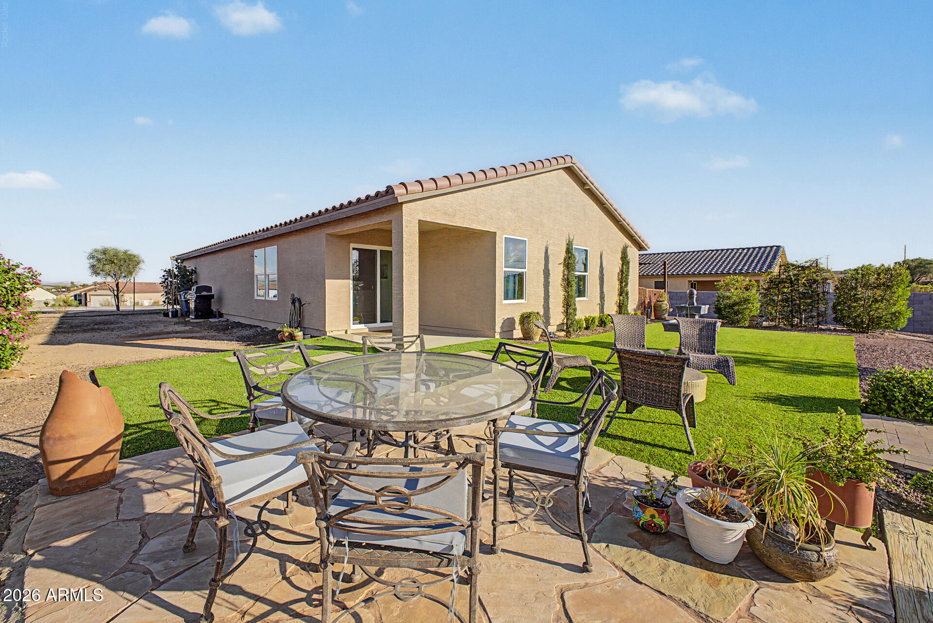 Home’s rear patio with a covered seating area, outdoor dining set, potted plants, and green lawn.