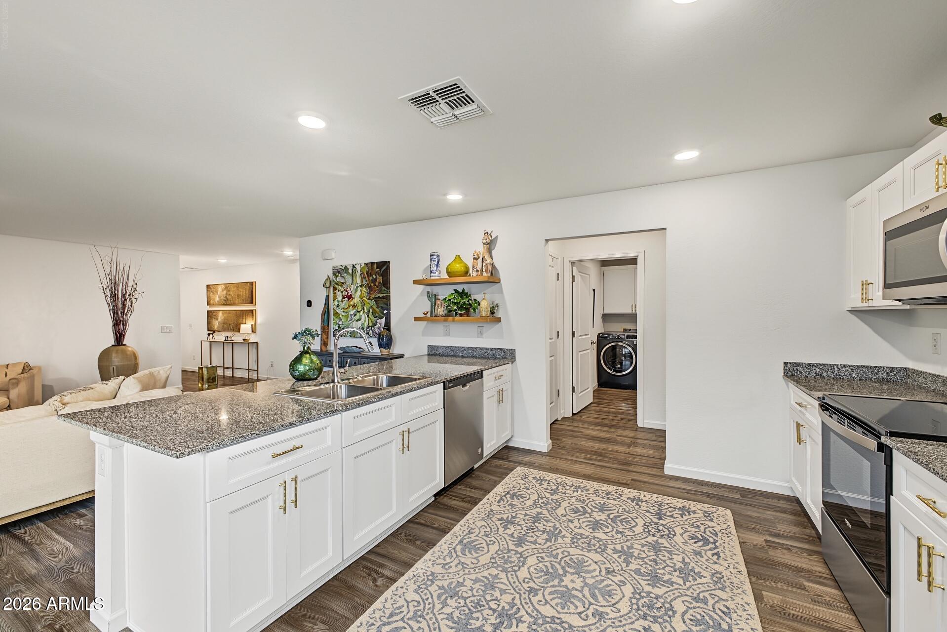 Open-plan kitchen with white cabinets, a granite island and double sink, stainless steel appliances, and a view into the living area.