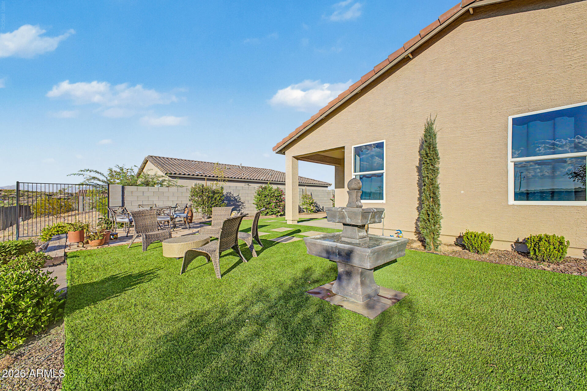 Landscaped backyard with a stone fountain, green lawn, and furnished patio beside a tan stucco home.