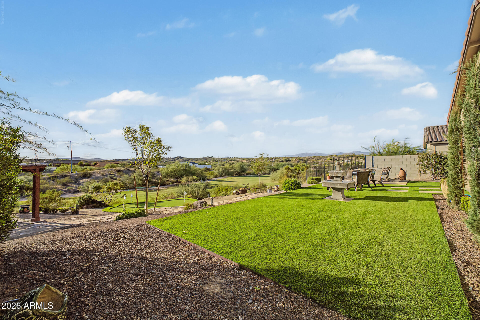 Backyard lawn and putting green with a stone fountain, outdoor dining area, and desert hills beyond.