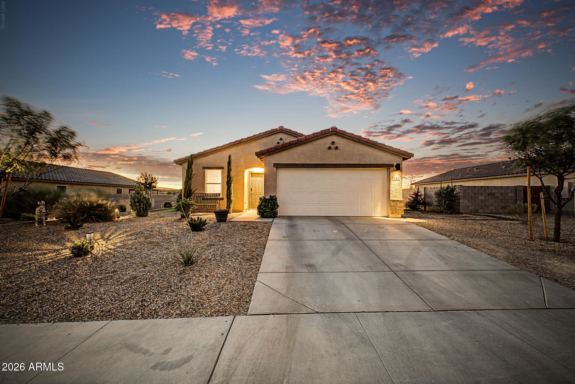 Single-story home with a two-car garage and desert landscaping under a colorful sunset sky.