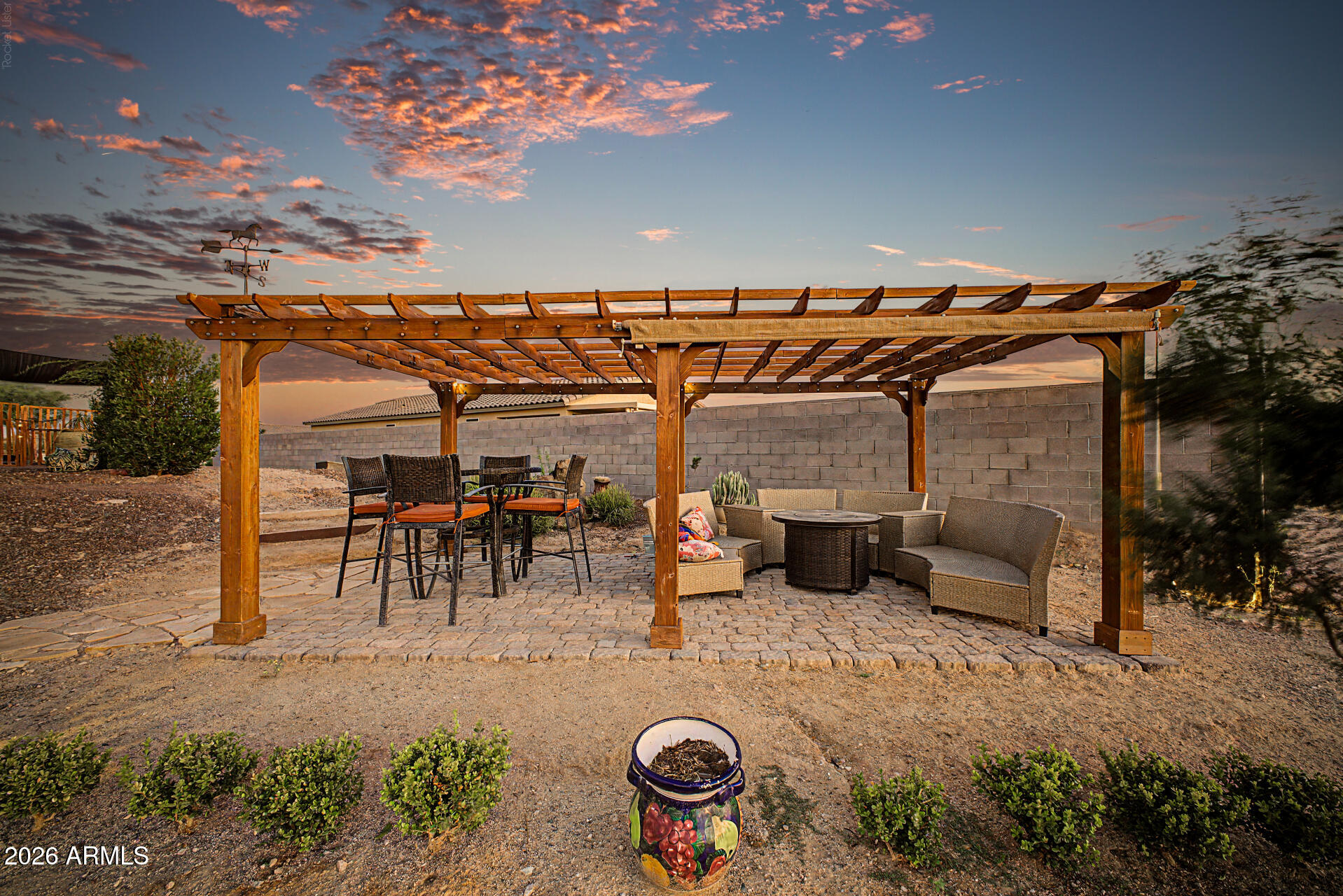 Backyard patio with a wooden pergola, outdoor dining and lounge seating, and a colorful sunset sky.