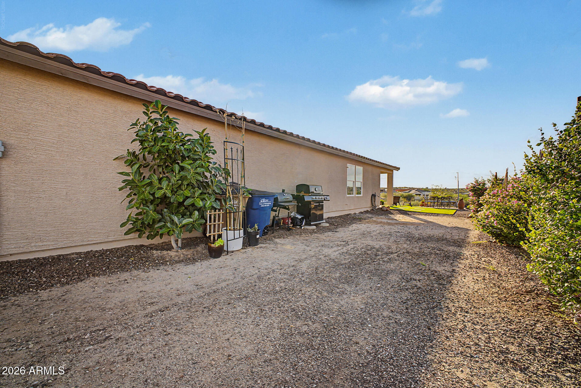 Side yard beside a stucco home, with a gravel path, grill, plants and a patio seating area.