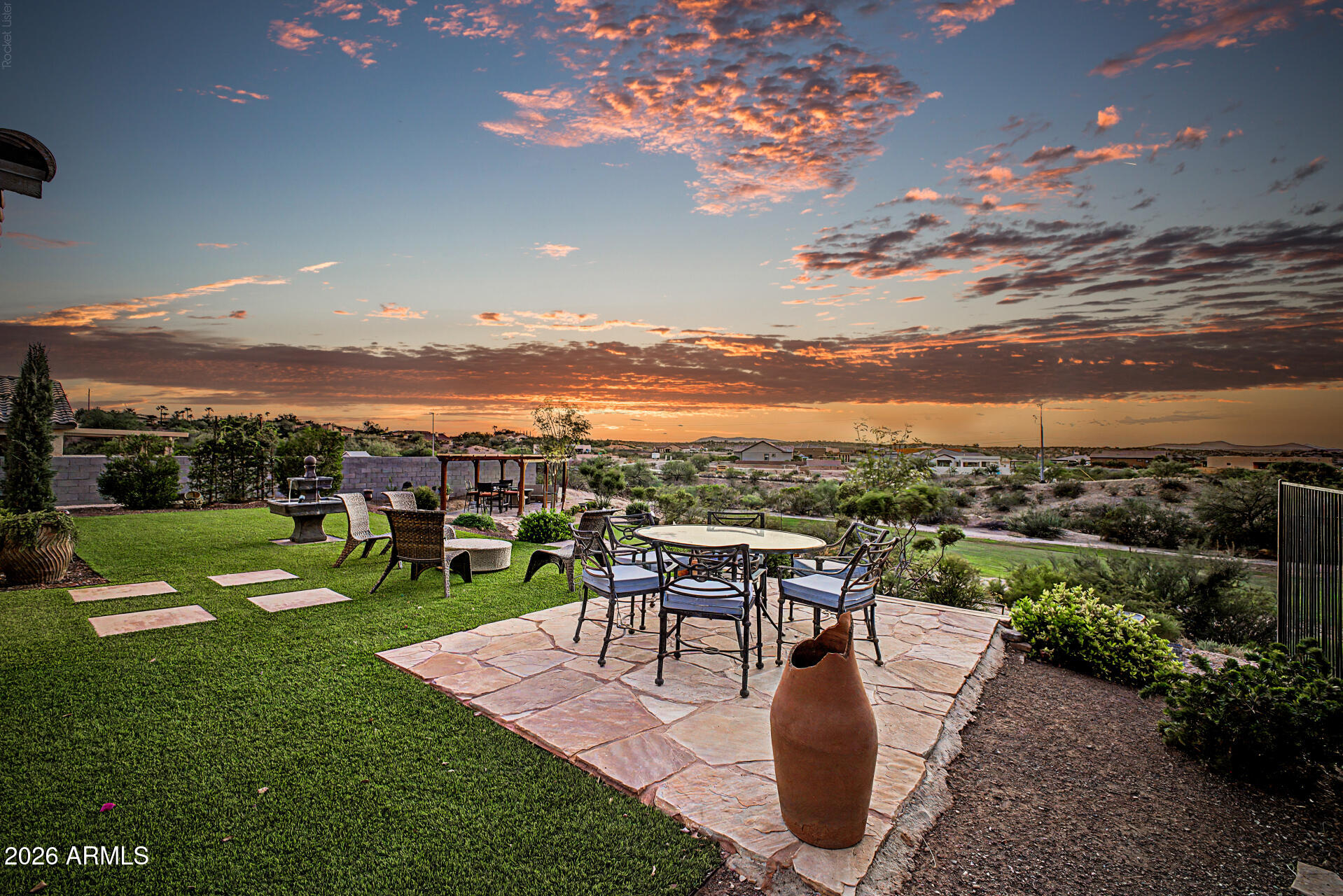 Furnished stone patio and lawn overlook a desert neighborhood beneath a vivid orange sunset.