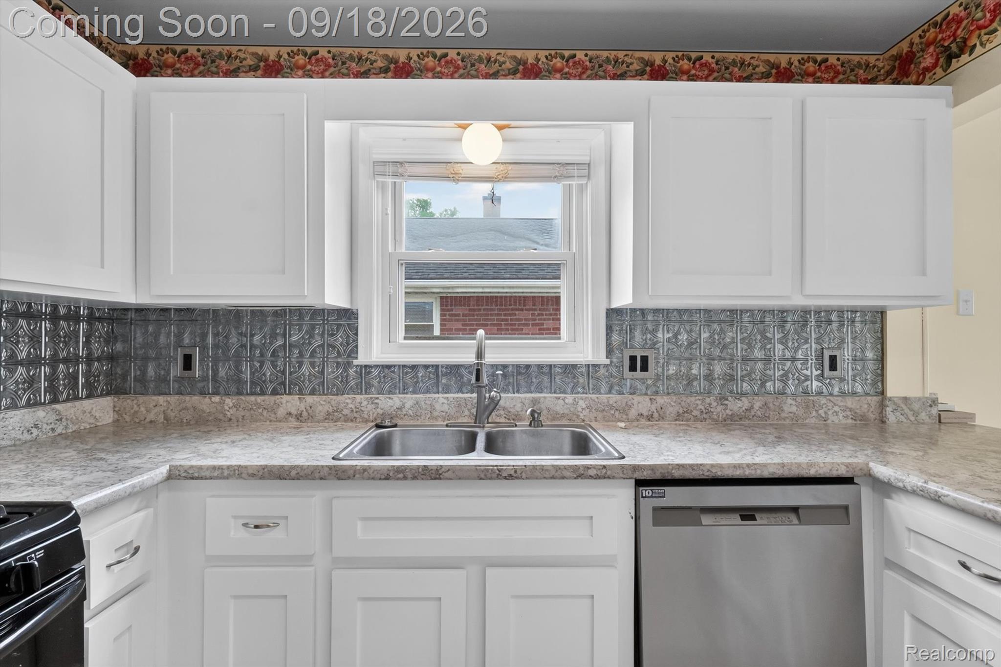 Kitchen sink beneath a window, with white cabinets, patterned gray backsplash, and stone countertops.