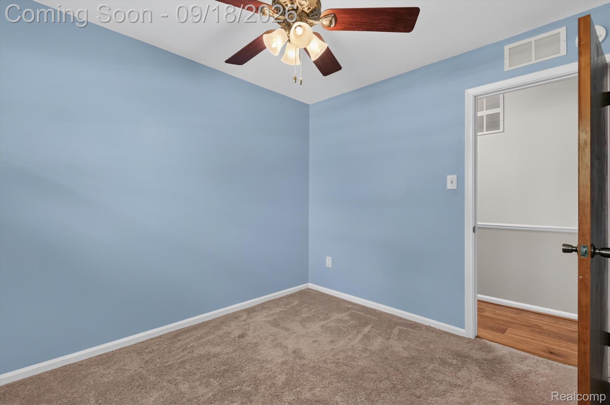 Empty bedroom with blue walls, beige carpet, and a ceiling fan; an open door reveals hardwood flooring.