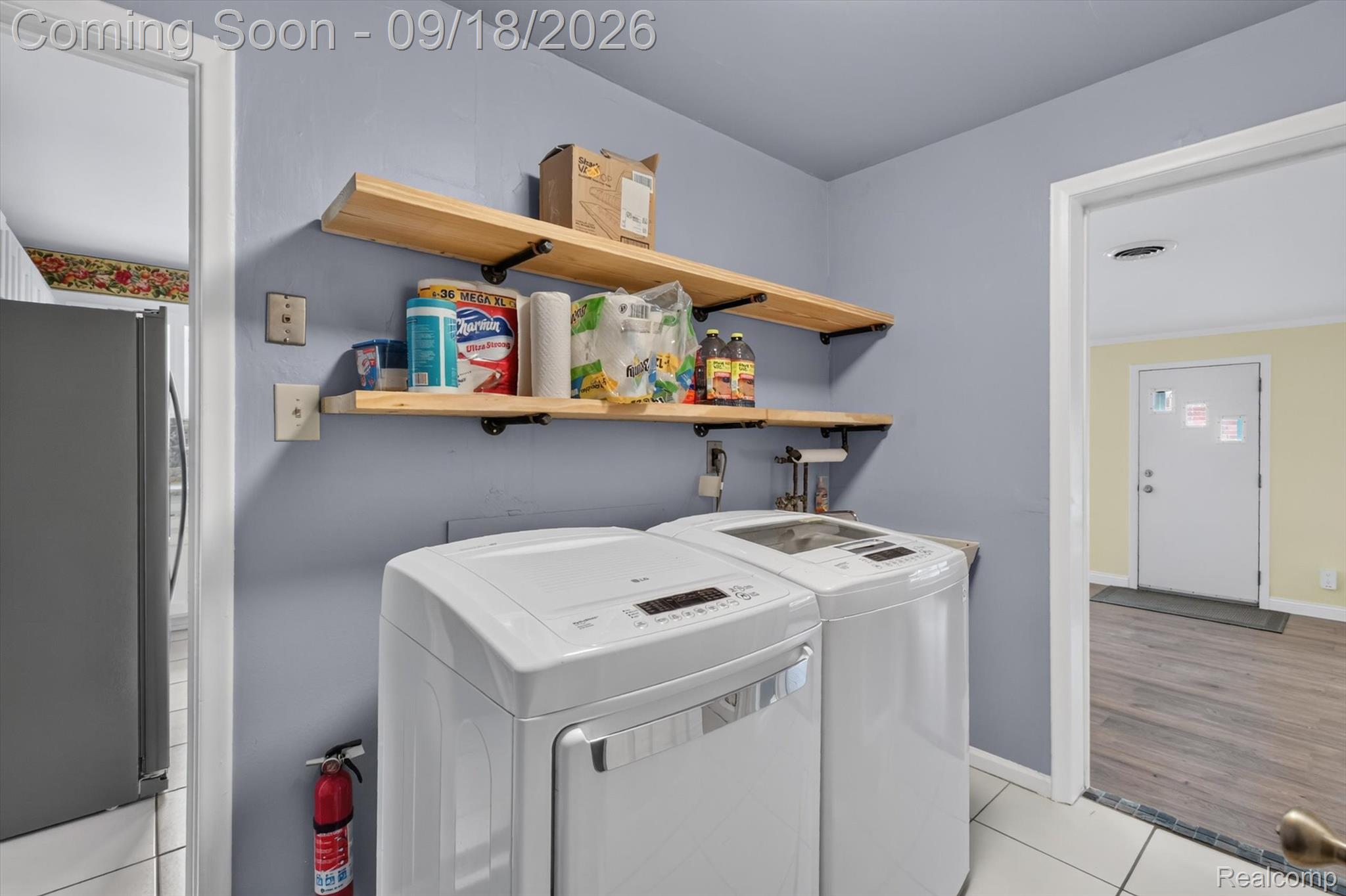 Laundry room with two white top-loading machines and wooden storage shelves.