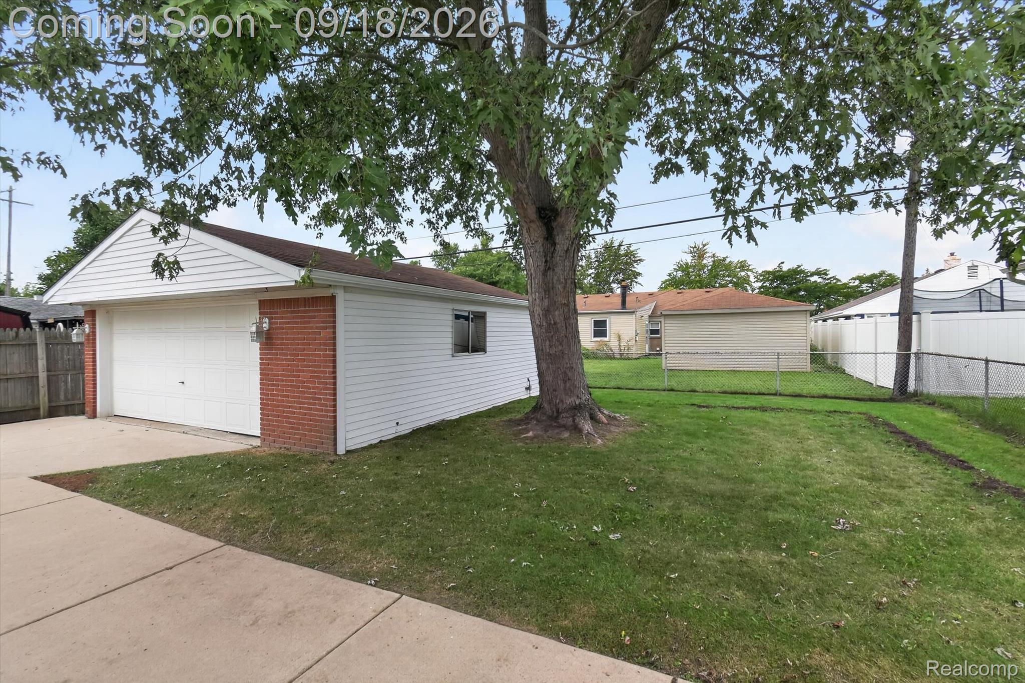 Detached white-sided garage beside a grassy fenced yard, shaded by a large tree.