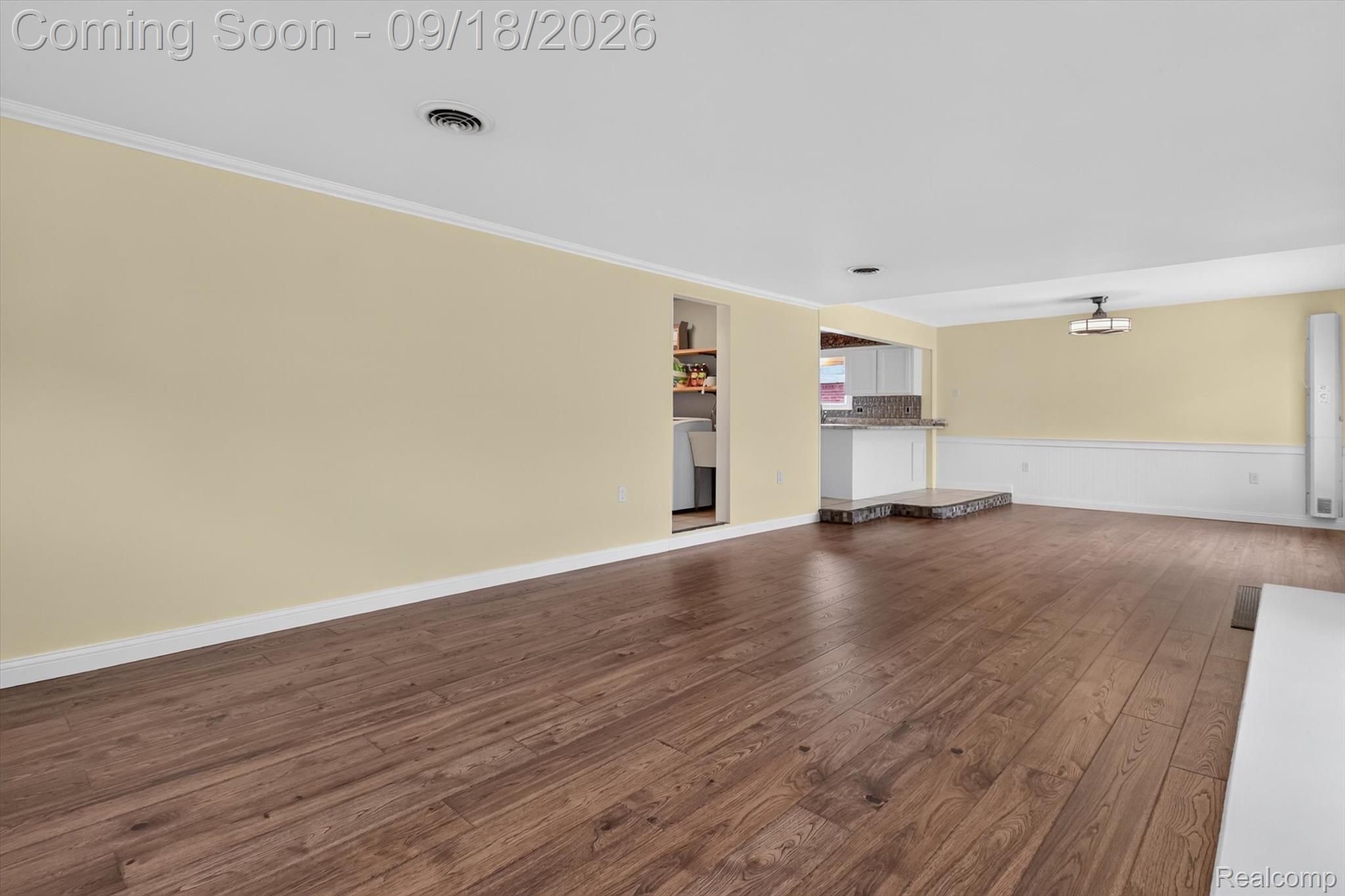 Mostly empty living area with wood plank floors, pale yellow walls, and an opening to the kitchen.