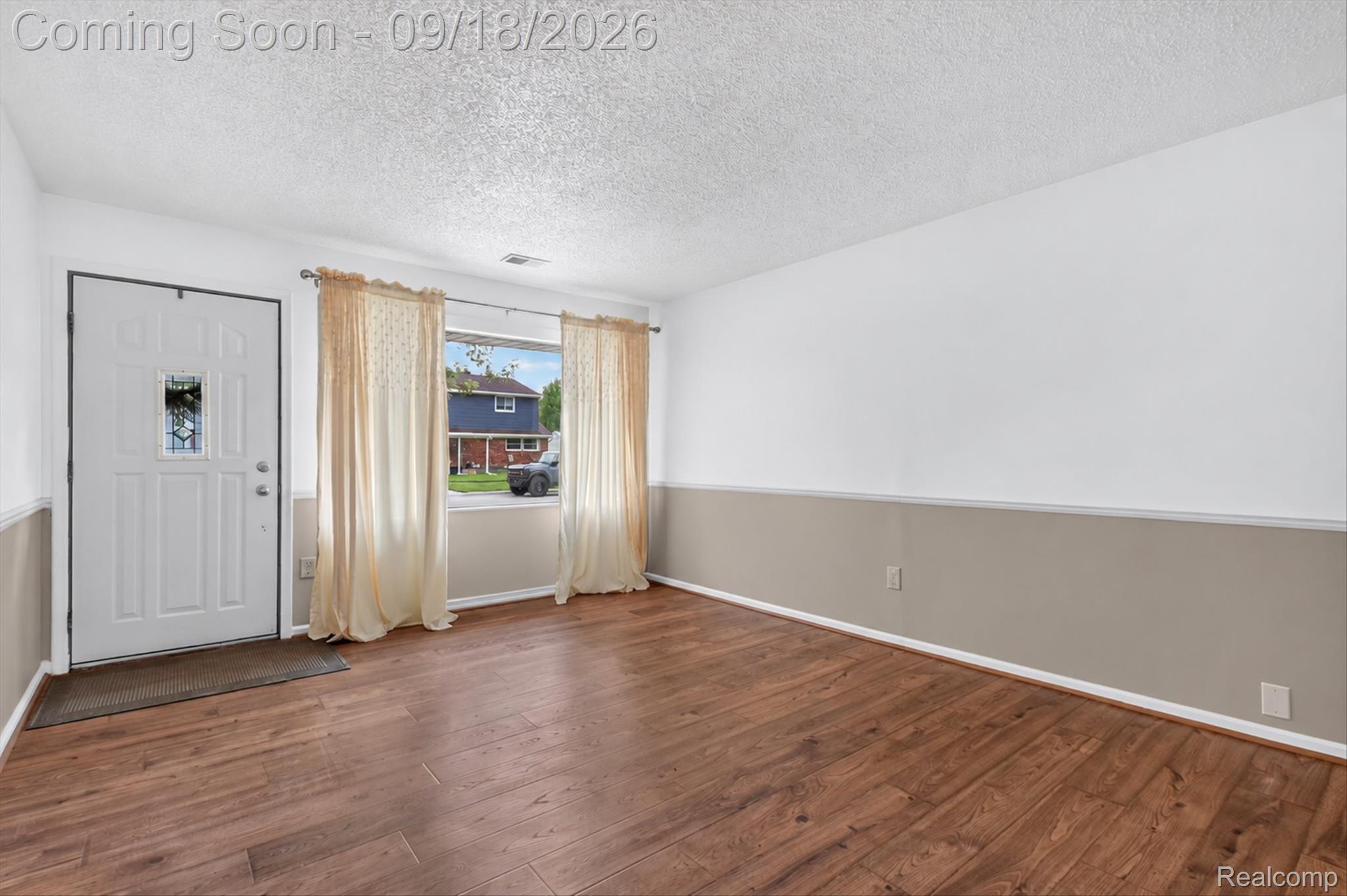 Vacant front room with wood-look flooring, a decorative-glass entry door, and a curtained window.