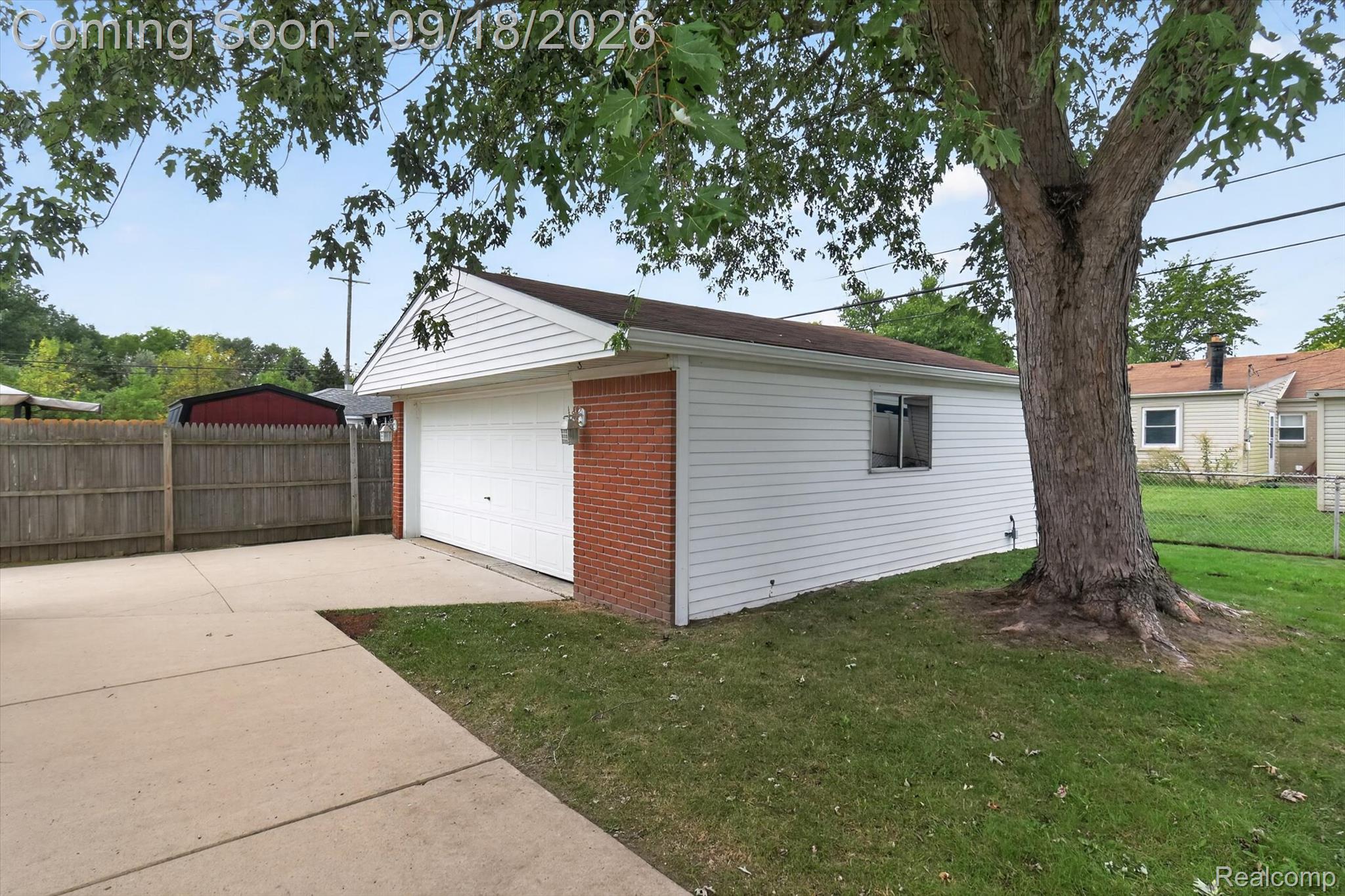 Detached white-sided garage with a brick front, concrete driveway, wooden fence, and large shade tree.