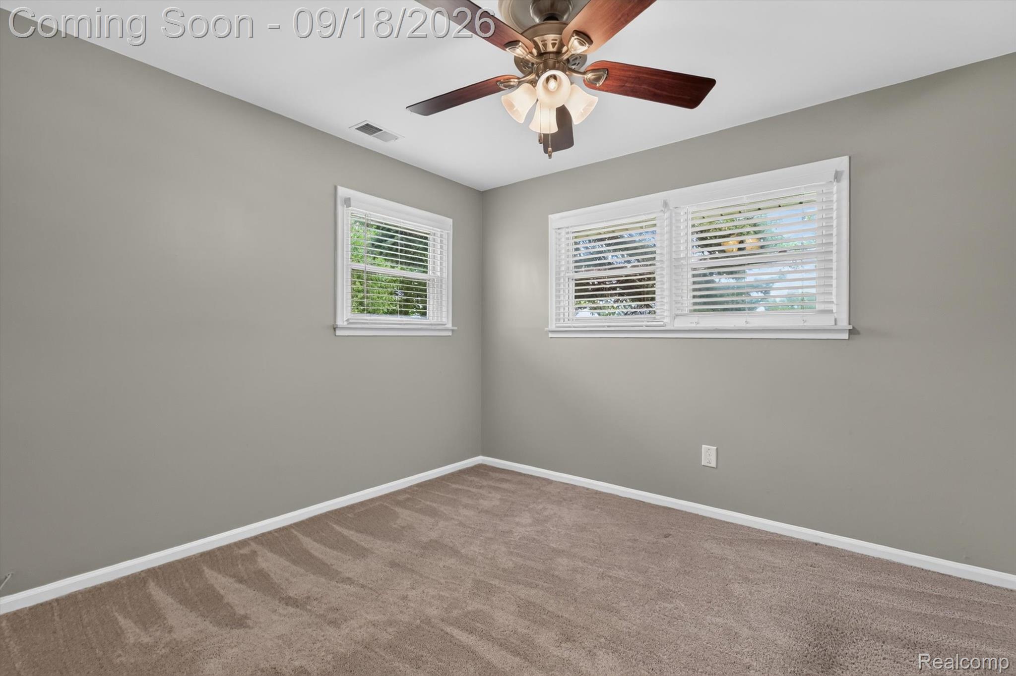 Empty bedroom with beige carpet, gray walls, two blinded windows, and a ceiling fan.