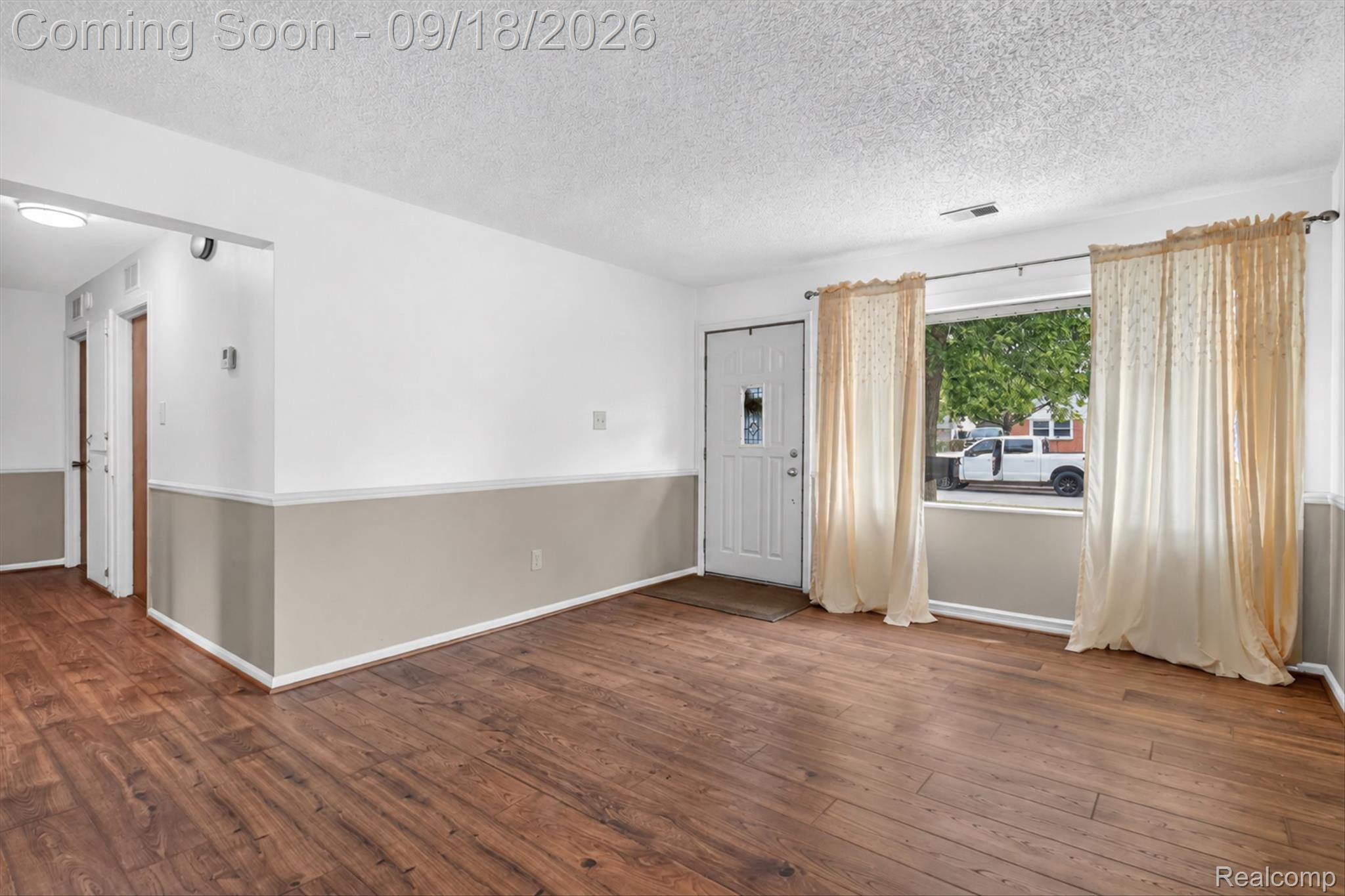 Empty living room with wood-look flooring, two-tone walls, and a curtained window beside the front door.