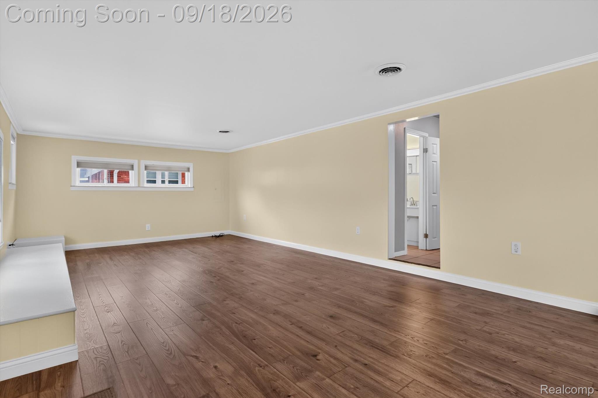 Empty room with wide-plank brown flooring, beige walls, high windows, and a doorway to a bathroom.