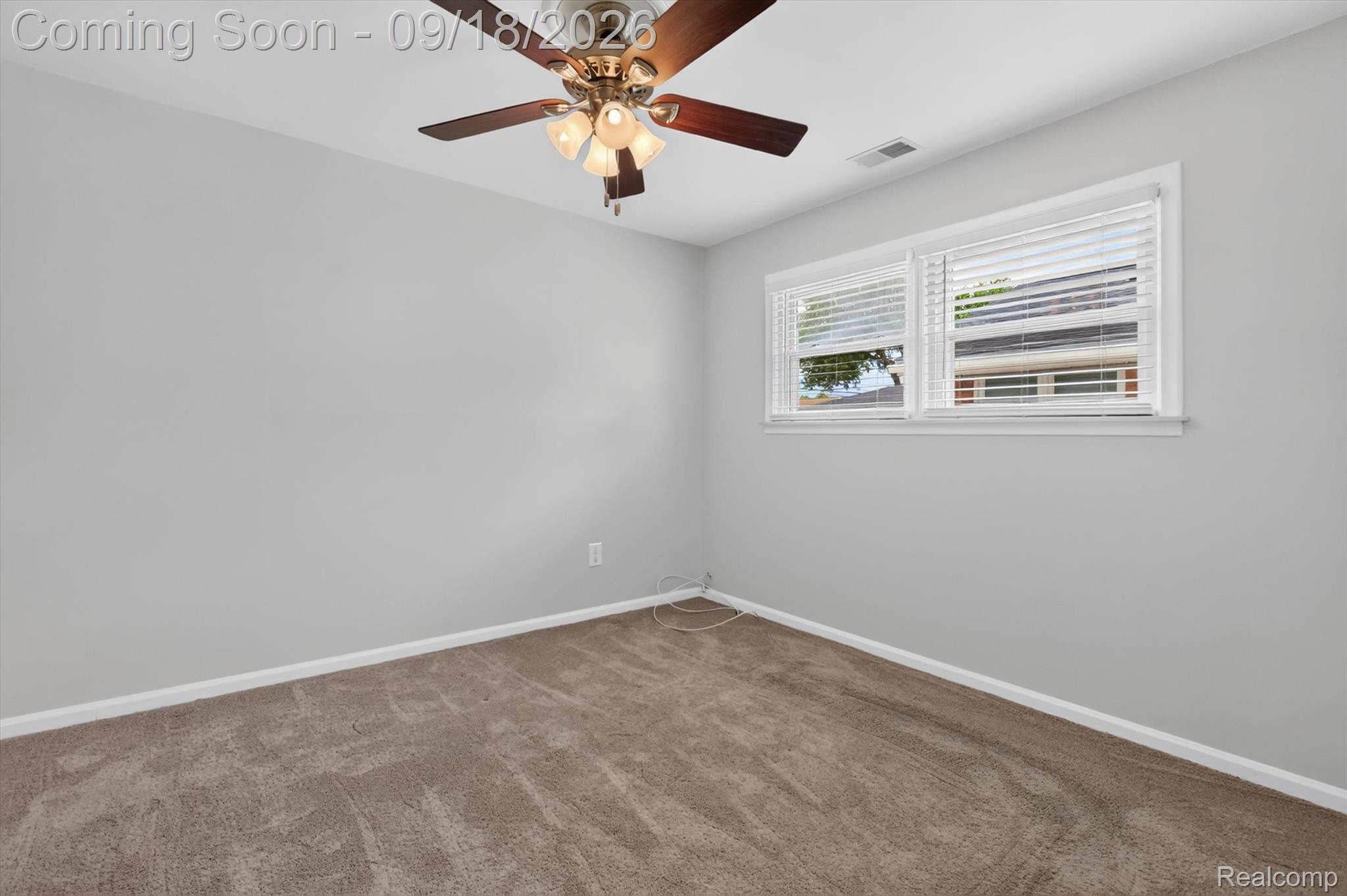 Empty bedroom with beige carpet, a lit ceiling fan, and two windows with white blinds.