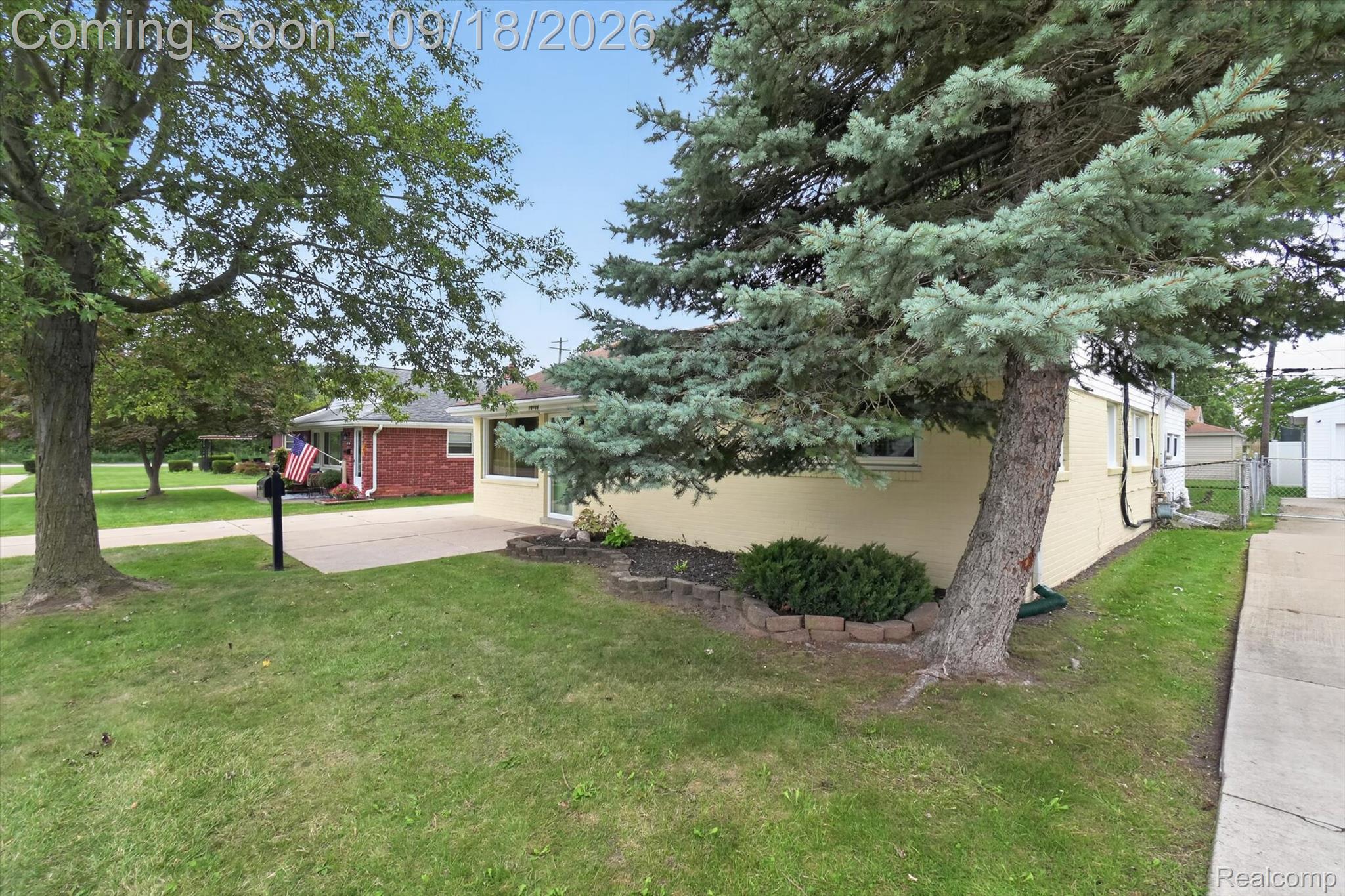 Single-story yellow-brick home partly screened by a large evergreen, with lawn and driveway.