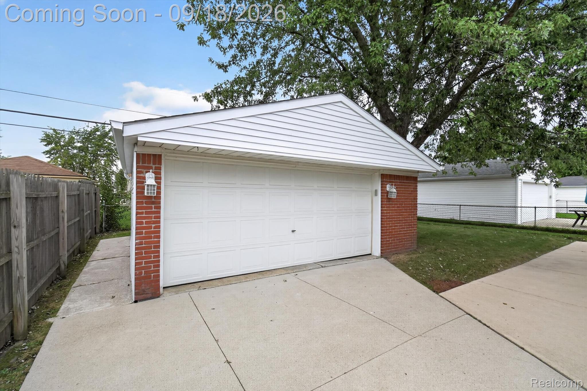 Garage with a wide white door, red brick supports, and a concrete driveway beneath a leafy tree.