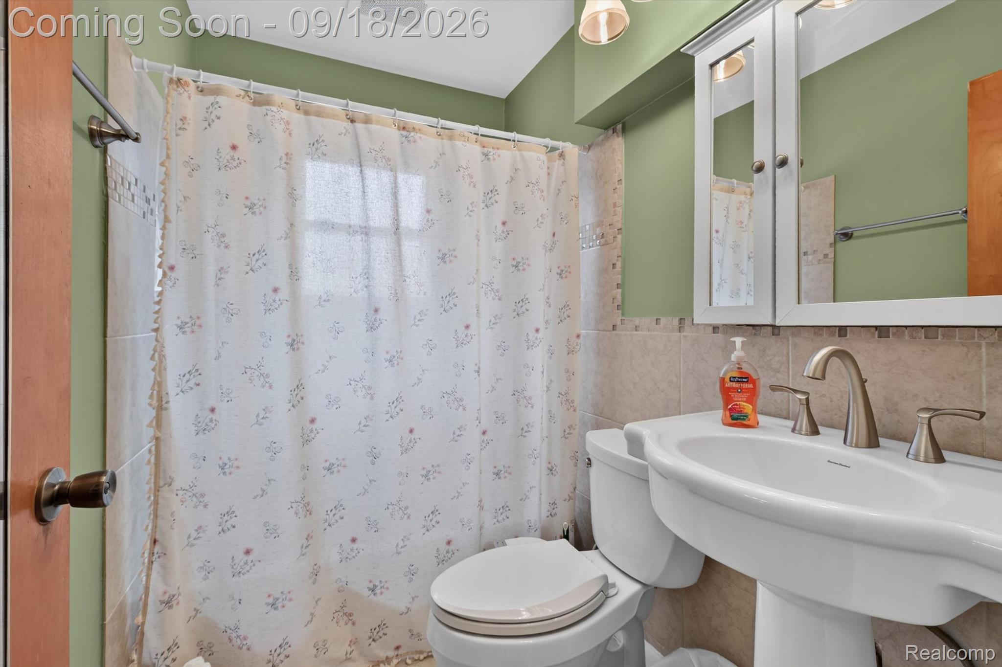 Bathroom with sage-green walls, floral shower curtain, mirrored cabinet, white toilet, and sink.