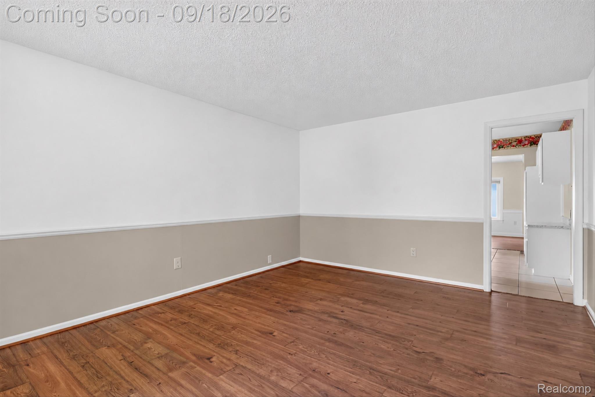 Vacant room with warm wood floors, two-tone walls, and a doorway to the kitchen.