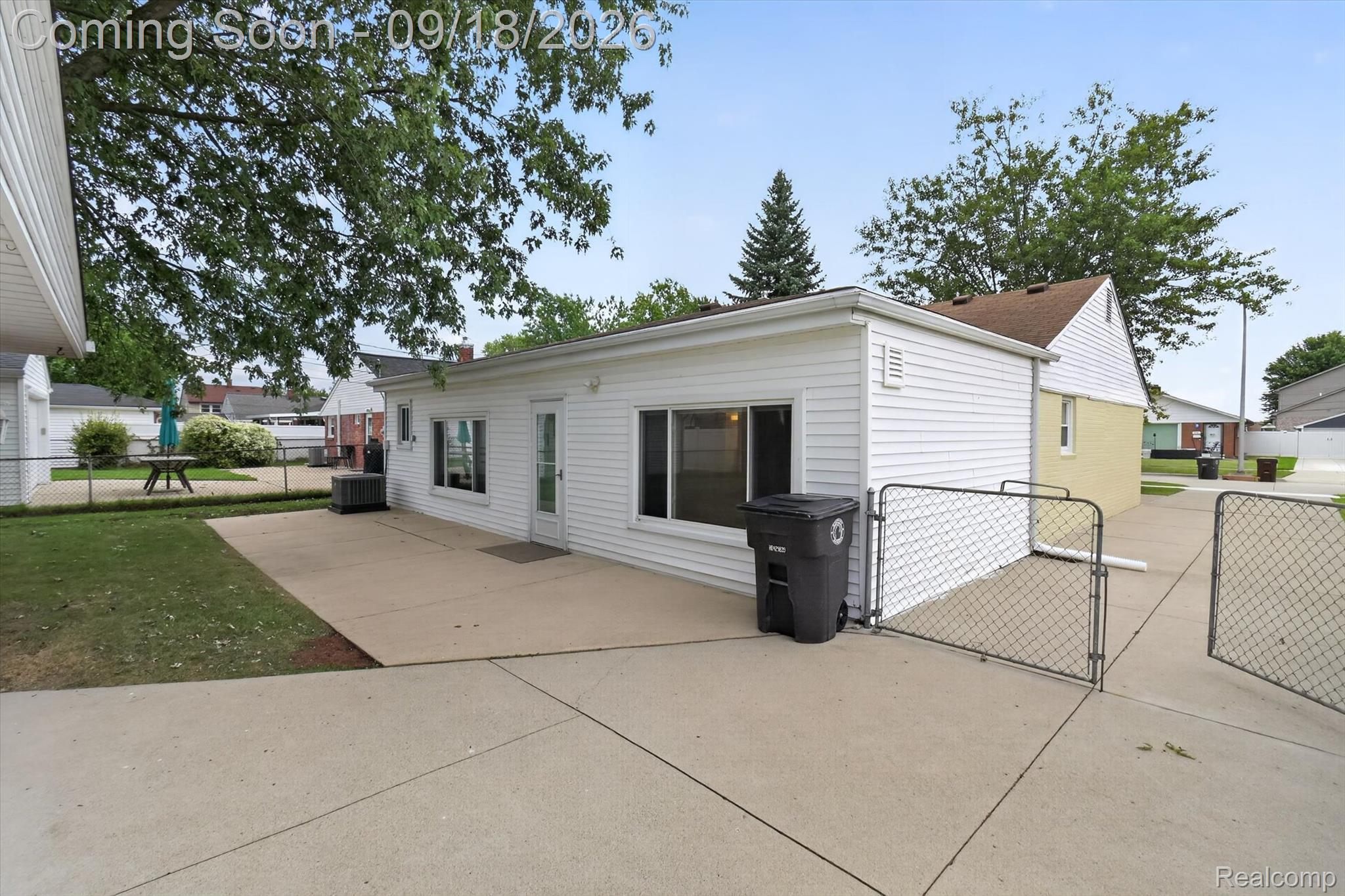 Rear of a white-sided house with a broad concrete patio and chain-link gates.