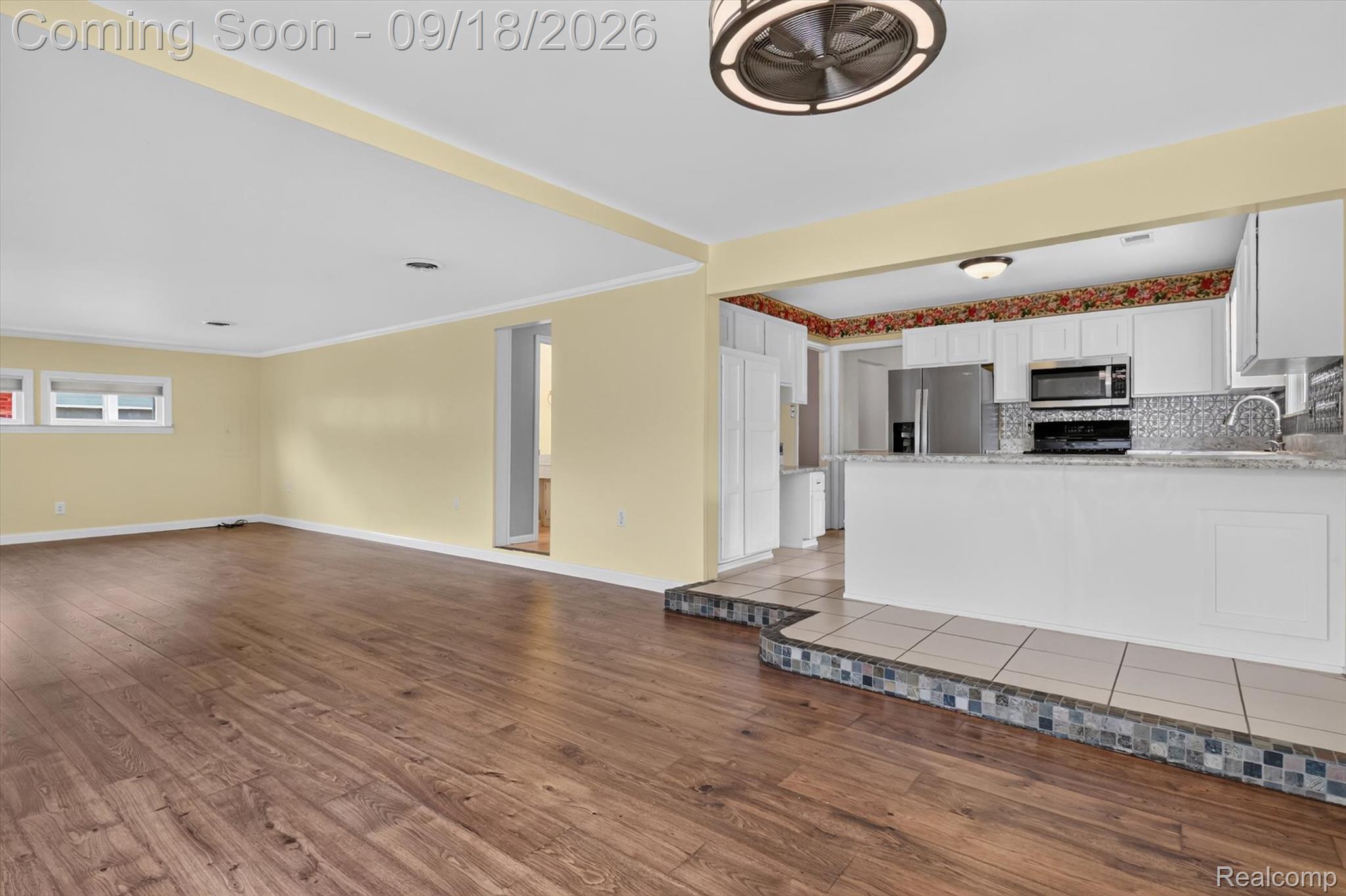 Open living area with wood flooring and adjoining white kitchen with stainless steel appliances.