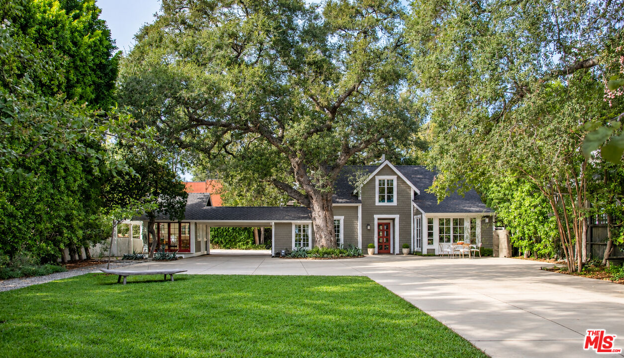 Gray house with a red front door beneath a sprawling tree, bordered by lawn and a wide driveway.