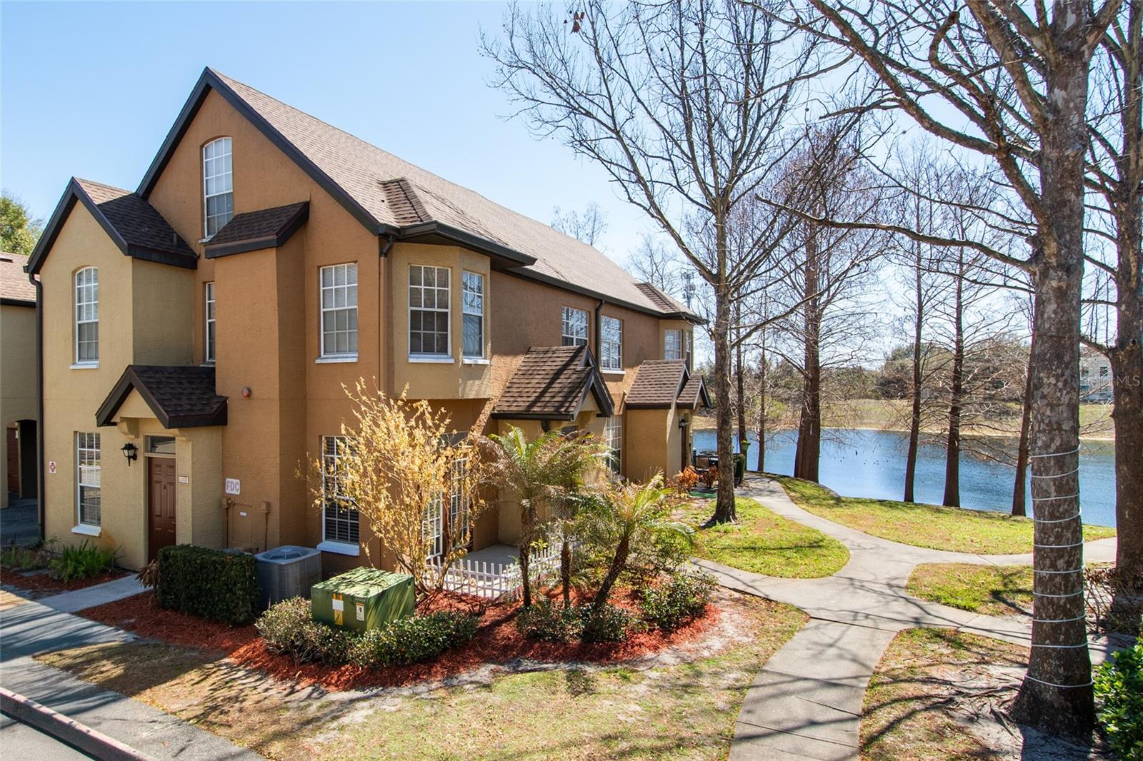Two-story tan residential building beside a lake, with a curved walkway along the waterfront.