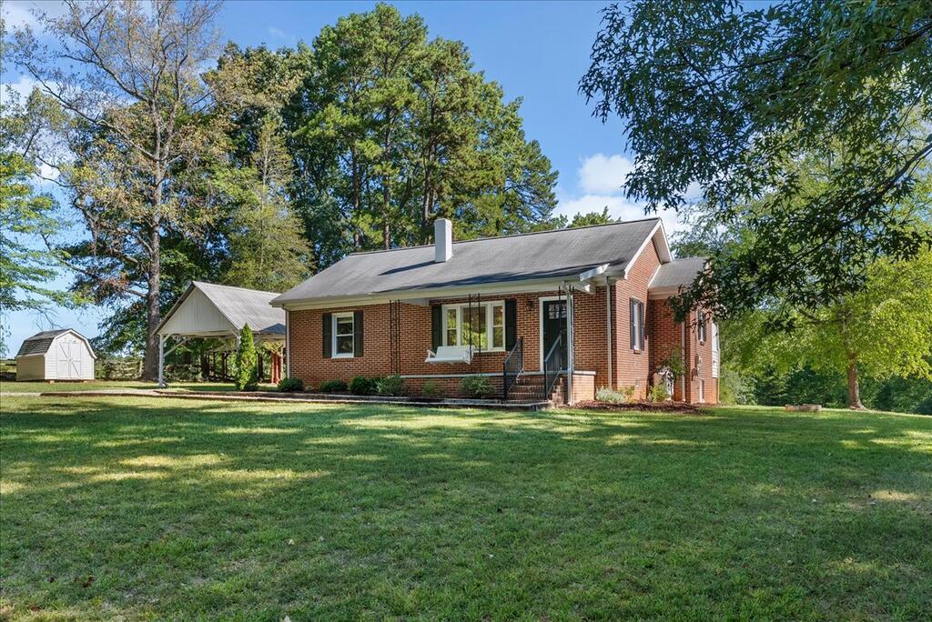 Single-story brick house with gray roof, front steps, broad lawn, shed, and carport.