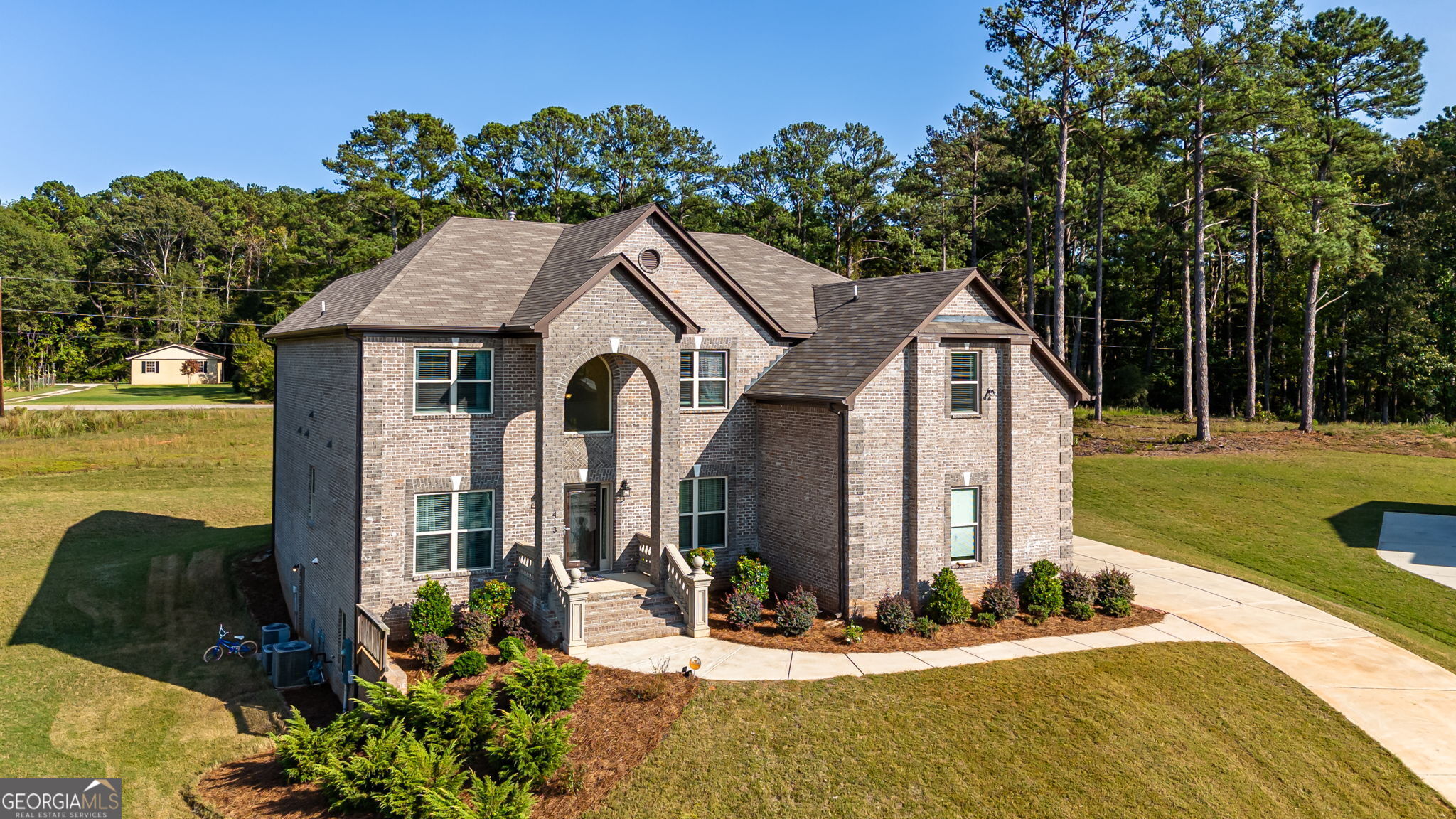 Two-story brick house with an arched entry, landscaped front beds, and a lawn bordered by pine trees.