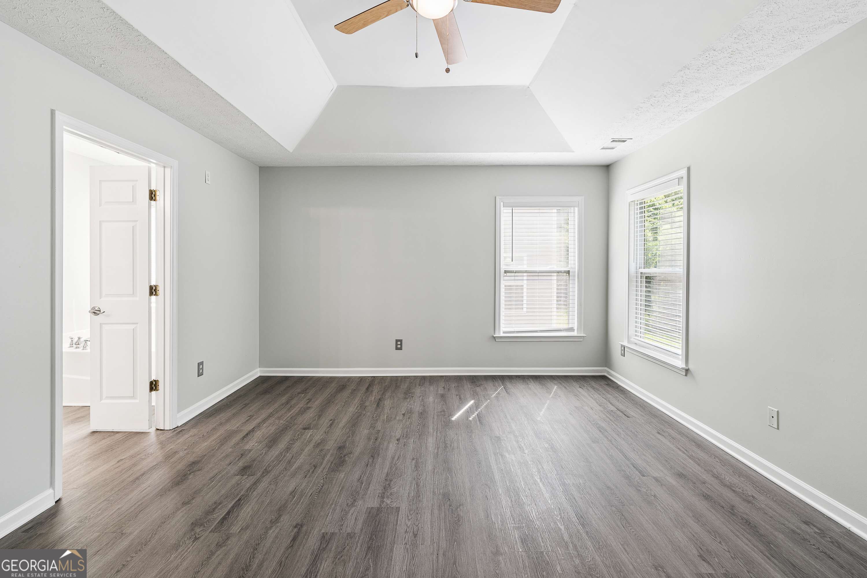 Empty bedroom with gray flooring, two window blinds, and a tray ceiling with ceiling fan.