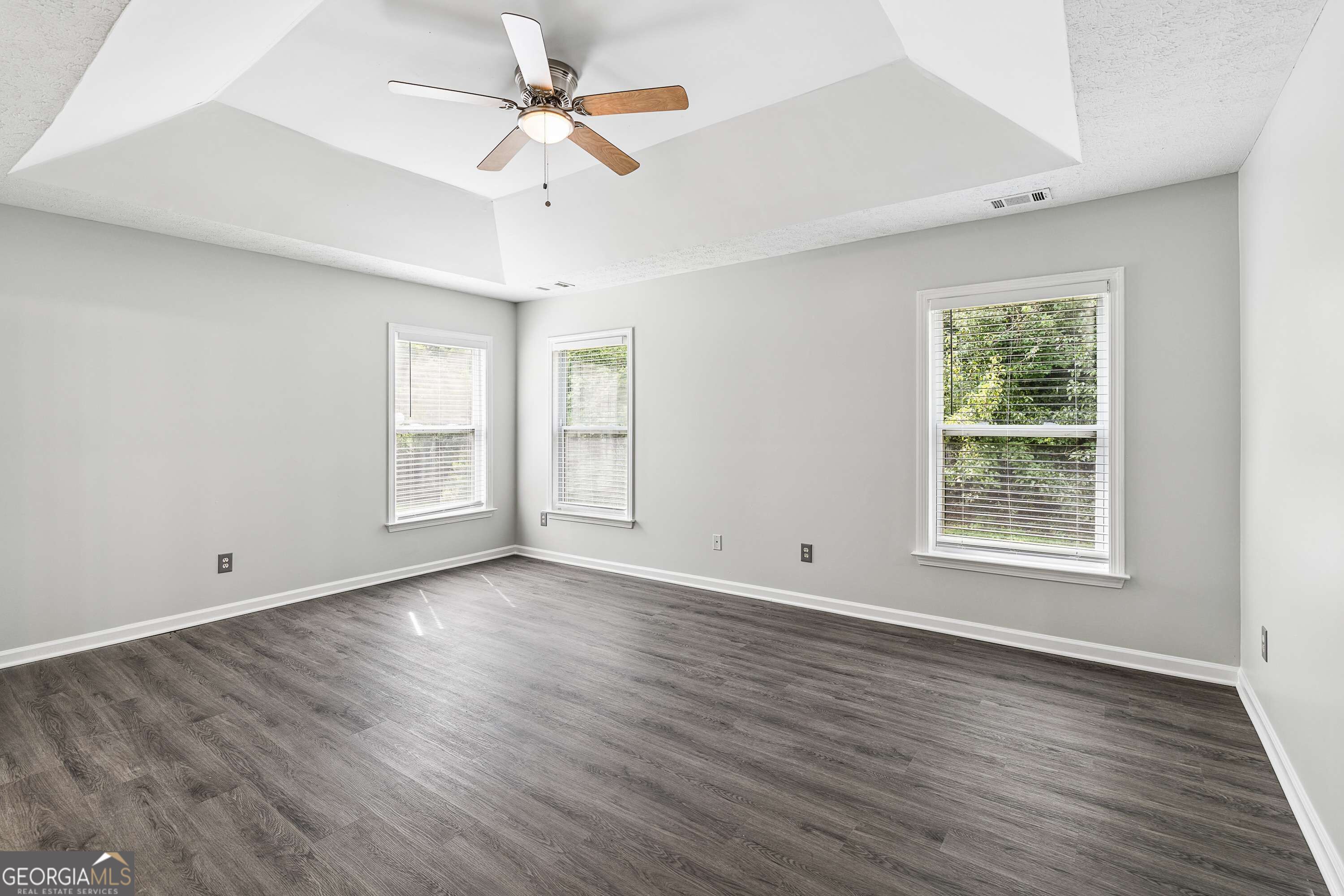 Vacant room with gray plank flooring, three windows with blinds, and a ceiling fan beneath a tray ceiling.