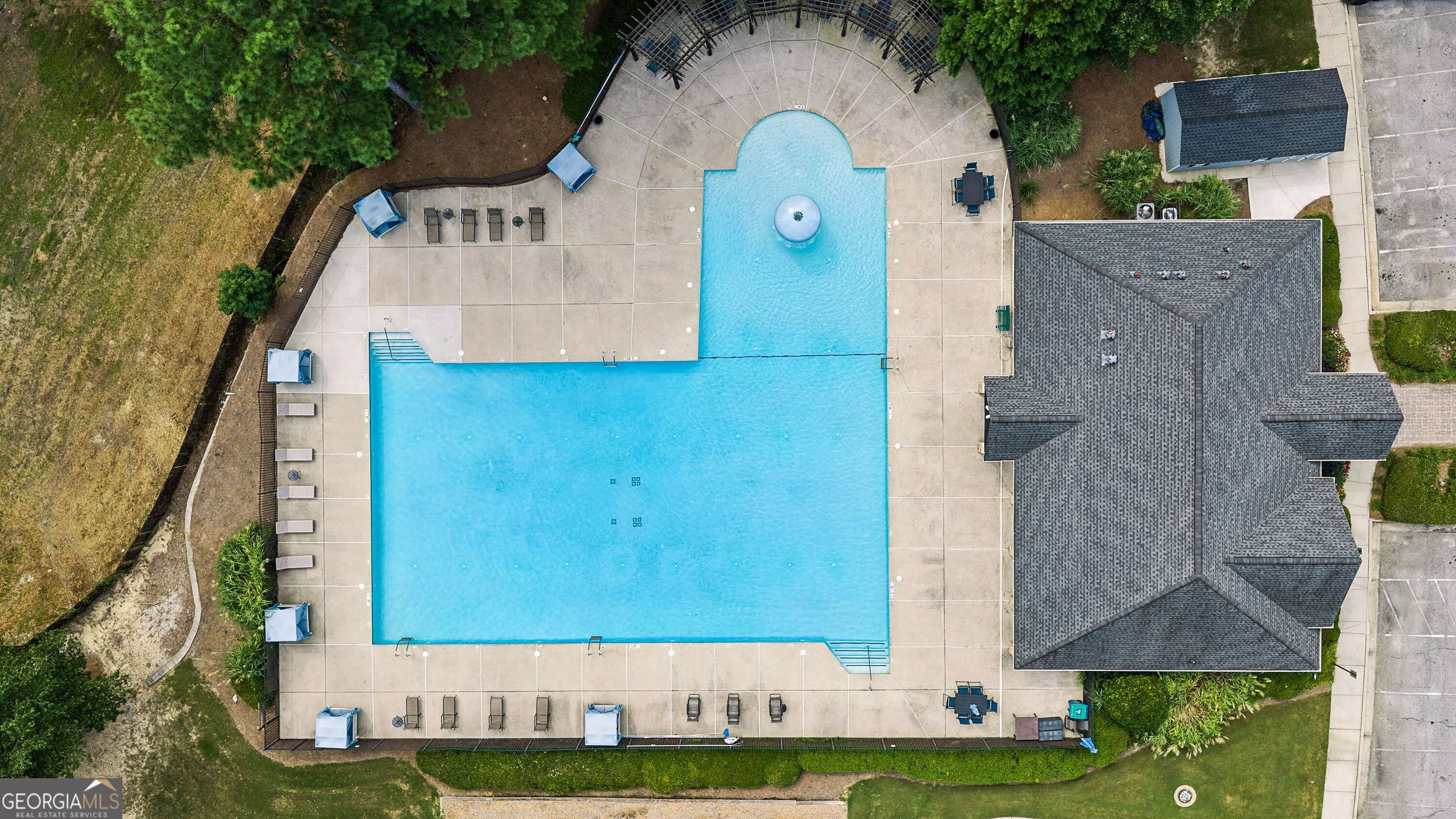 Aerial view of a large community pool with a fountain, paved deck, lounge chairs, and adjacent clubhouse.